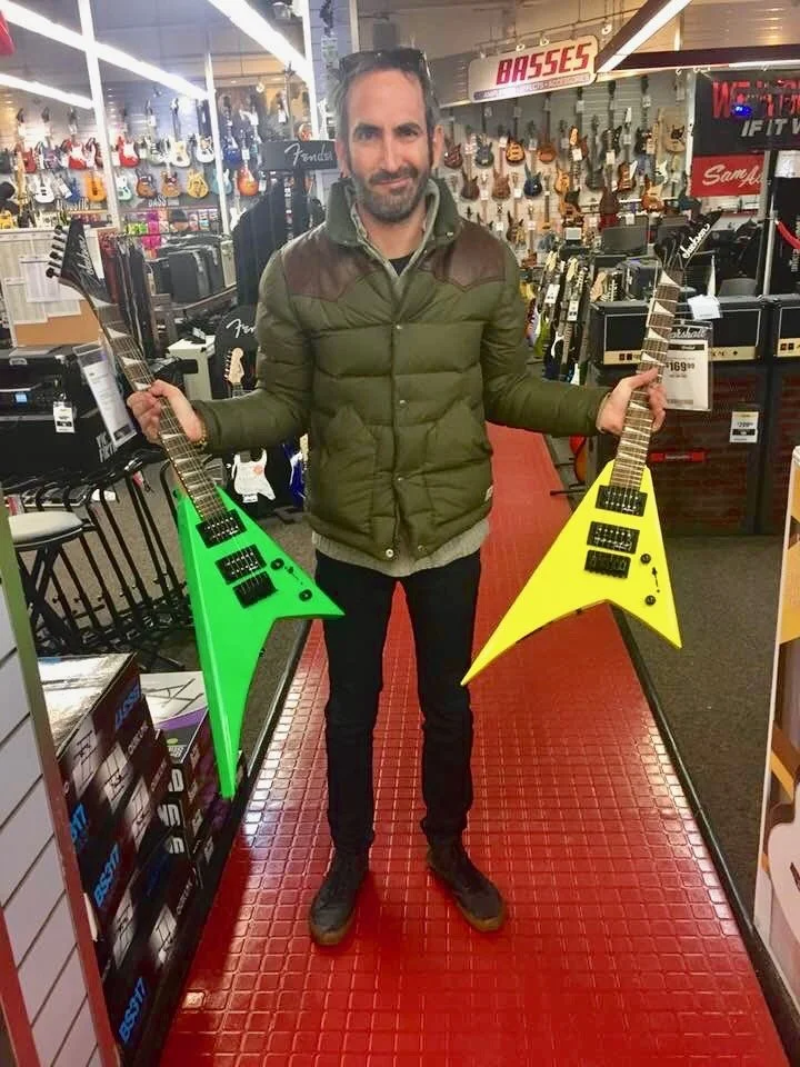A man standing in a music store holding two guitar-shaped objects, one neon green and one bright yellow, with a display of guitars and musical equipment behind him.
