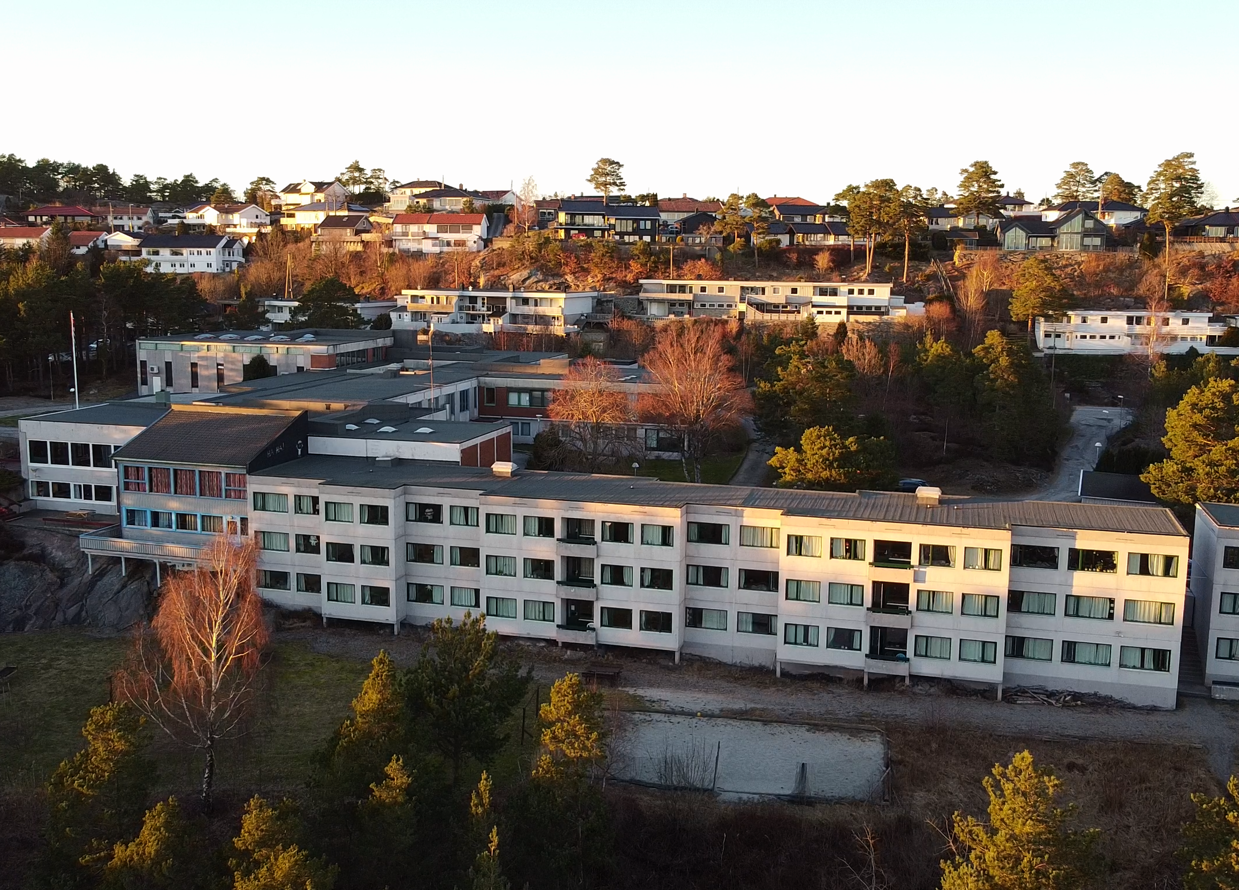 Aerial view of a hillside residential area with modern houses and apartment buildings surrounded by trees in the late afternoon sunlight.