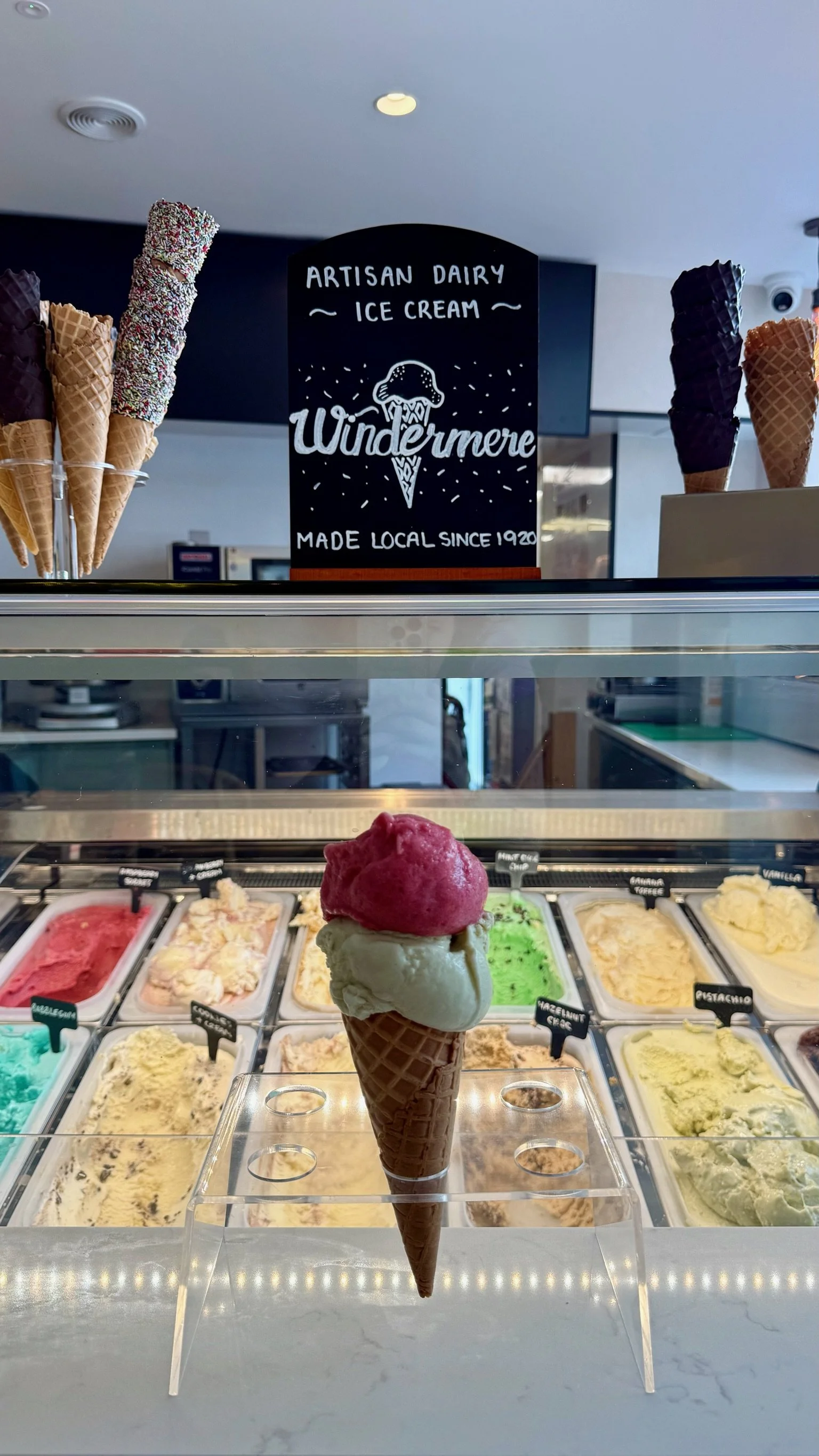 Ice cream shop display case with a triple scoop cone in the foreground and various flavors of ice cream in the background. Sign reads "Artisan Dairy Ice Cream Wondermere Made Local Since 1920."