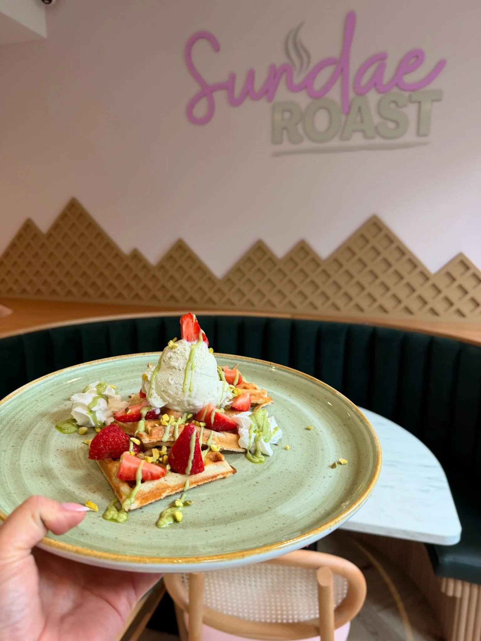 A plate with a waffle topped with strawberries, pistachios, whipped cream, and a scoop of ice cream, held by a hand in a cafe or restaurant setting.