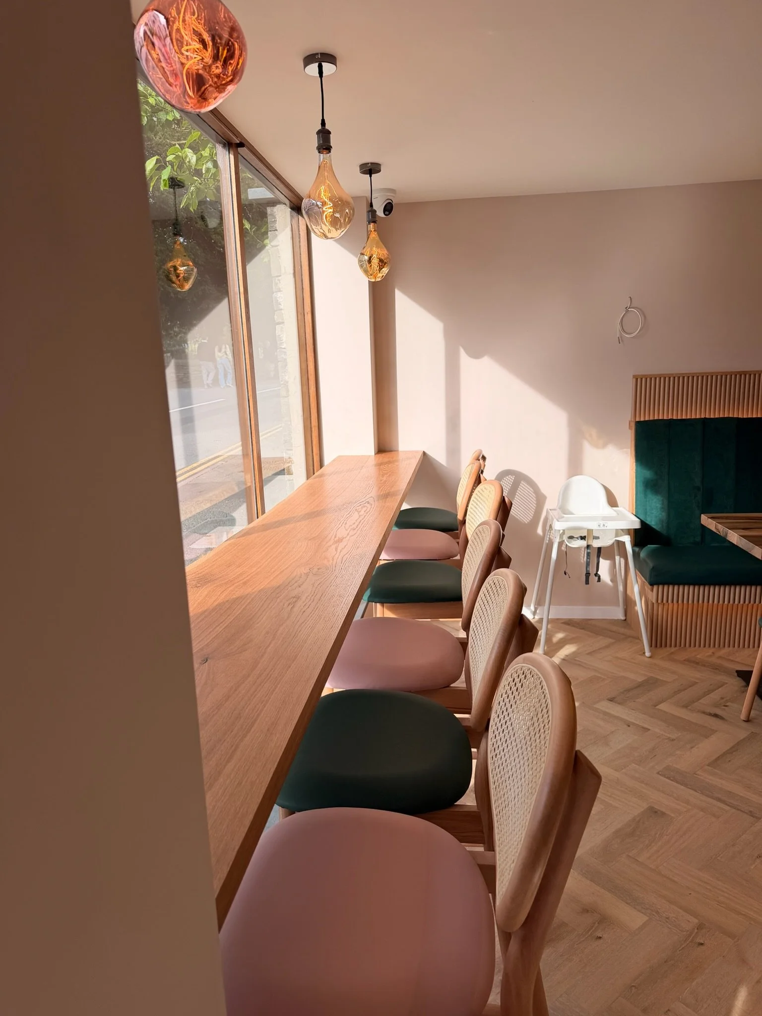 Interior of a cafe with a long wooden counter along glass windows, various colorful chairs, and hanging pendant lights, with a high chair and some details of the seating area visible.