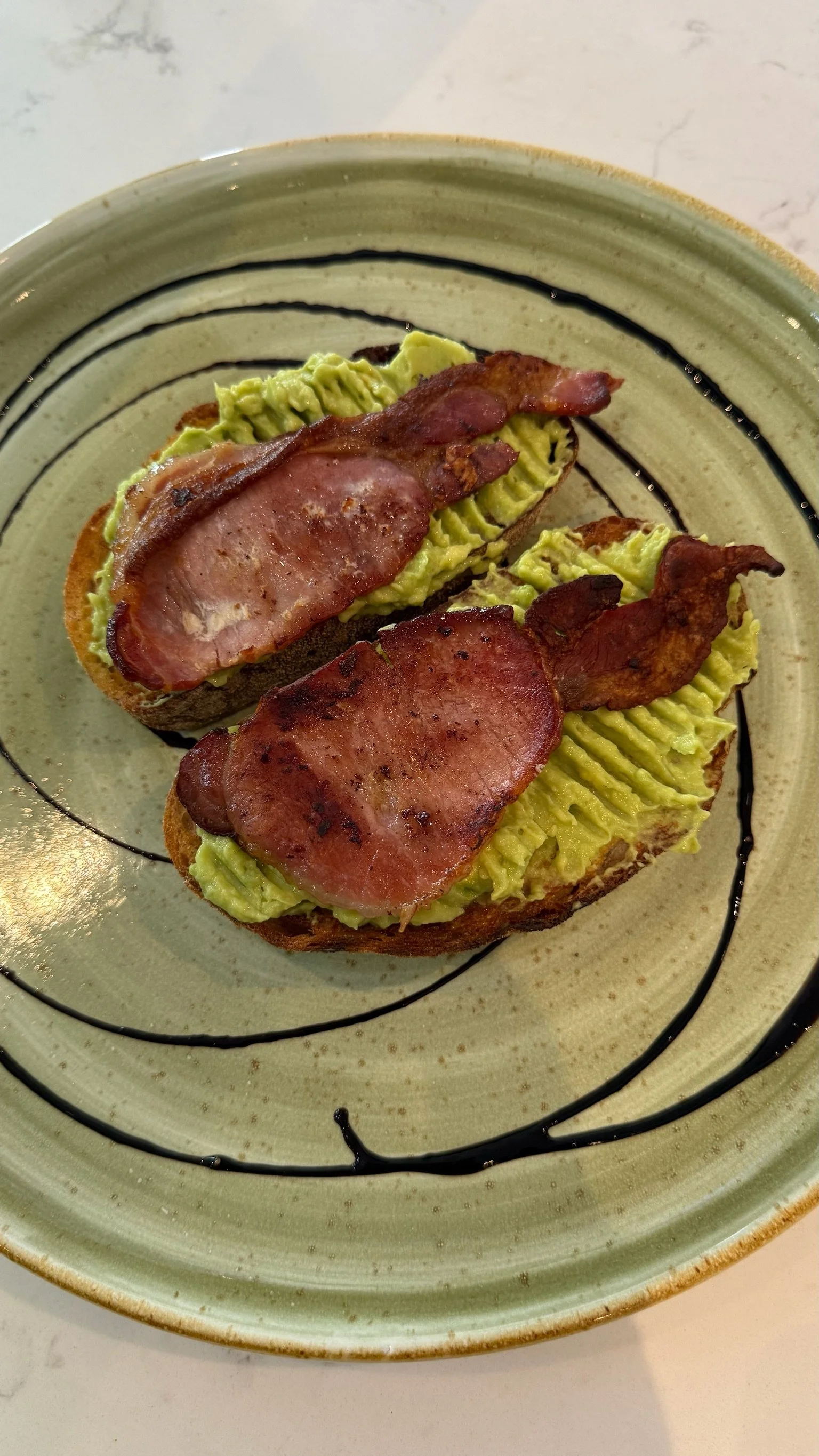 Slices of toasted bread topped with mashed avocado and crispy bacon slices, drizzled with balsamic glaze on a decorative plate.