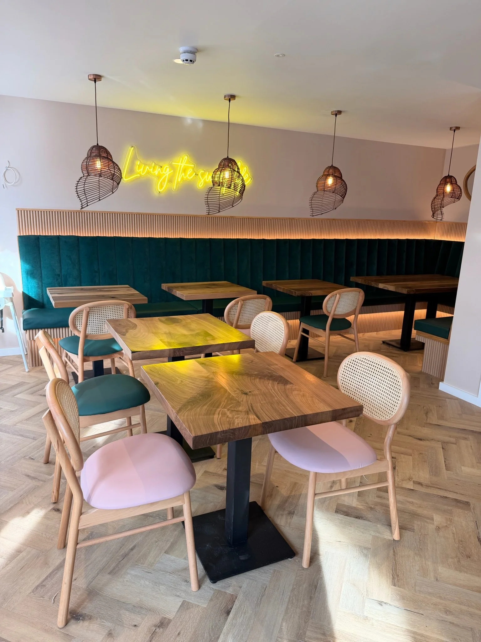 A cozy cafe dining area with wooden tables, pastel-colored chairs, a green cushioned banquette, and a wall with a yellow neon sign that reads 'Living the sun'. Hanging woven pendant lights add warm lighting to the space, which has a wooden floor.