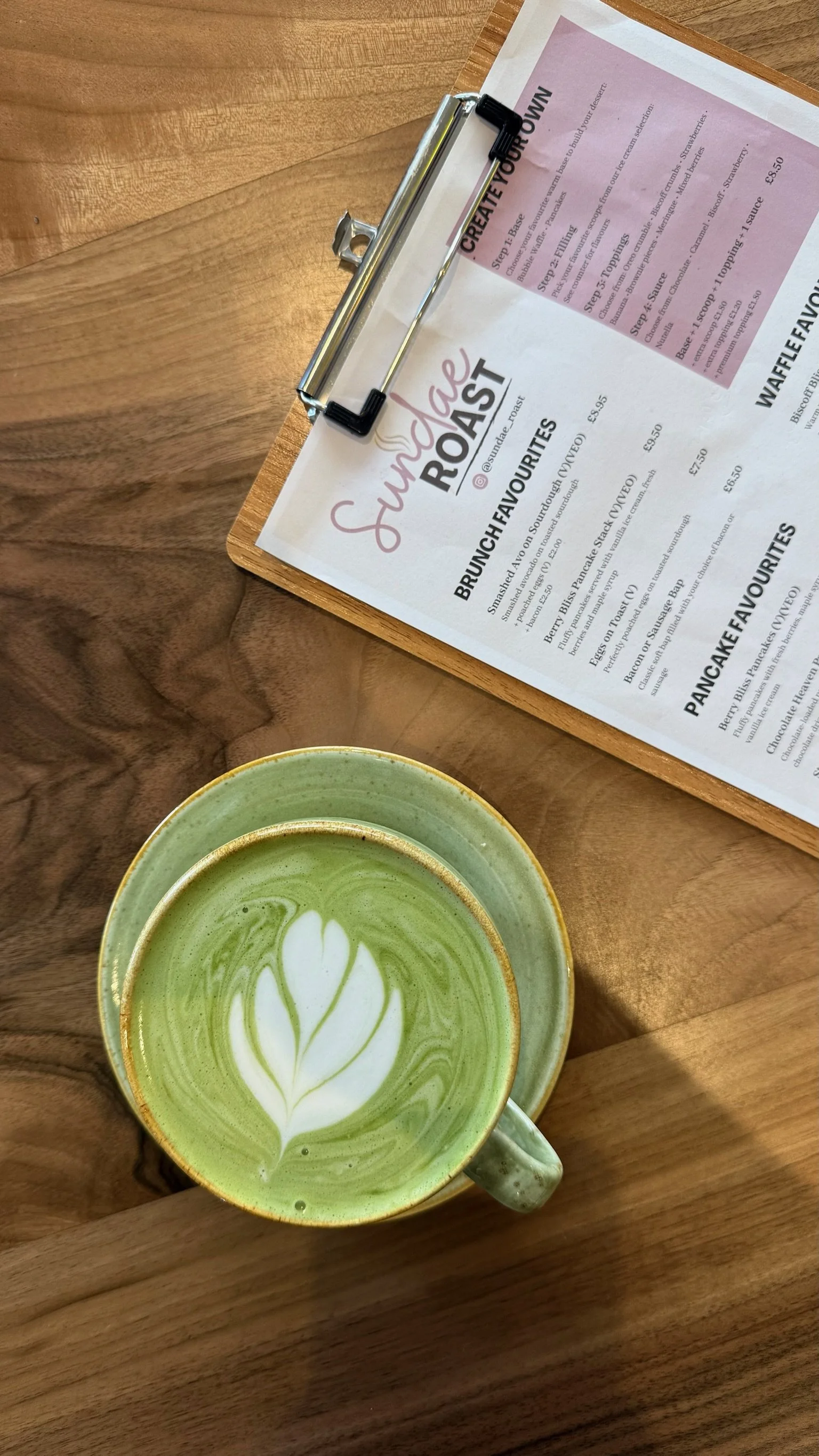 A green matcha latte with foam art in a ceramic mug on a wooden table, with a menu and a clipboard nearby.