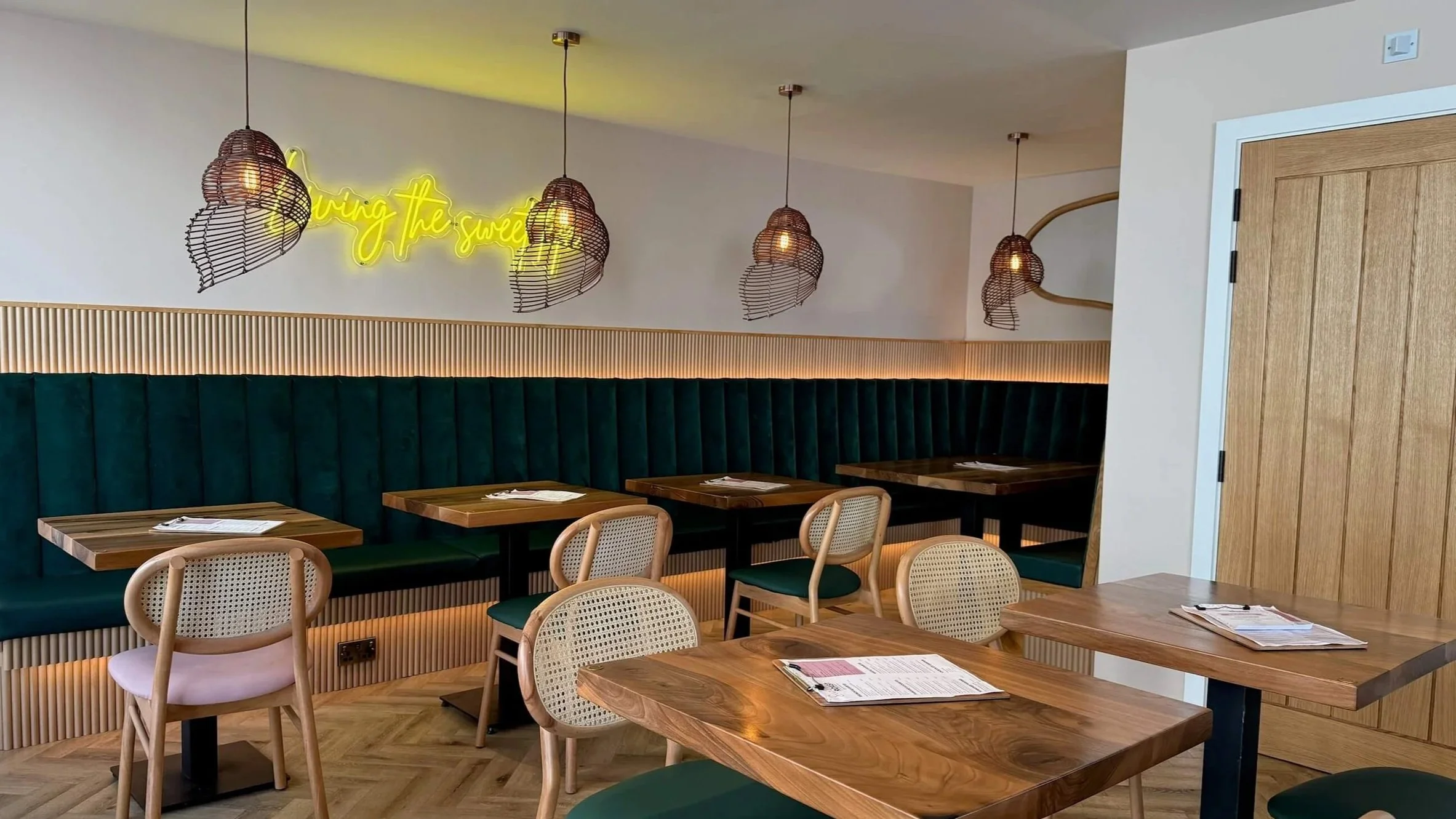 Interior of a modern restaurant with wooden tables, light-colored chairs, a dark green upholstered booth, hanging woven pendant lights, a wood door, and a yellow neon sign on the wall that says 'Living the sweet'.