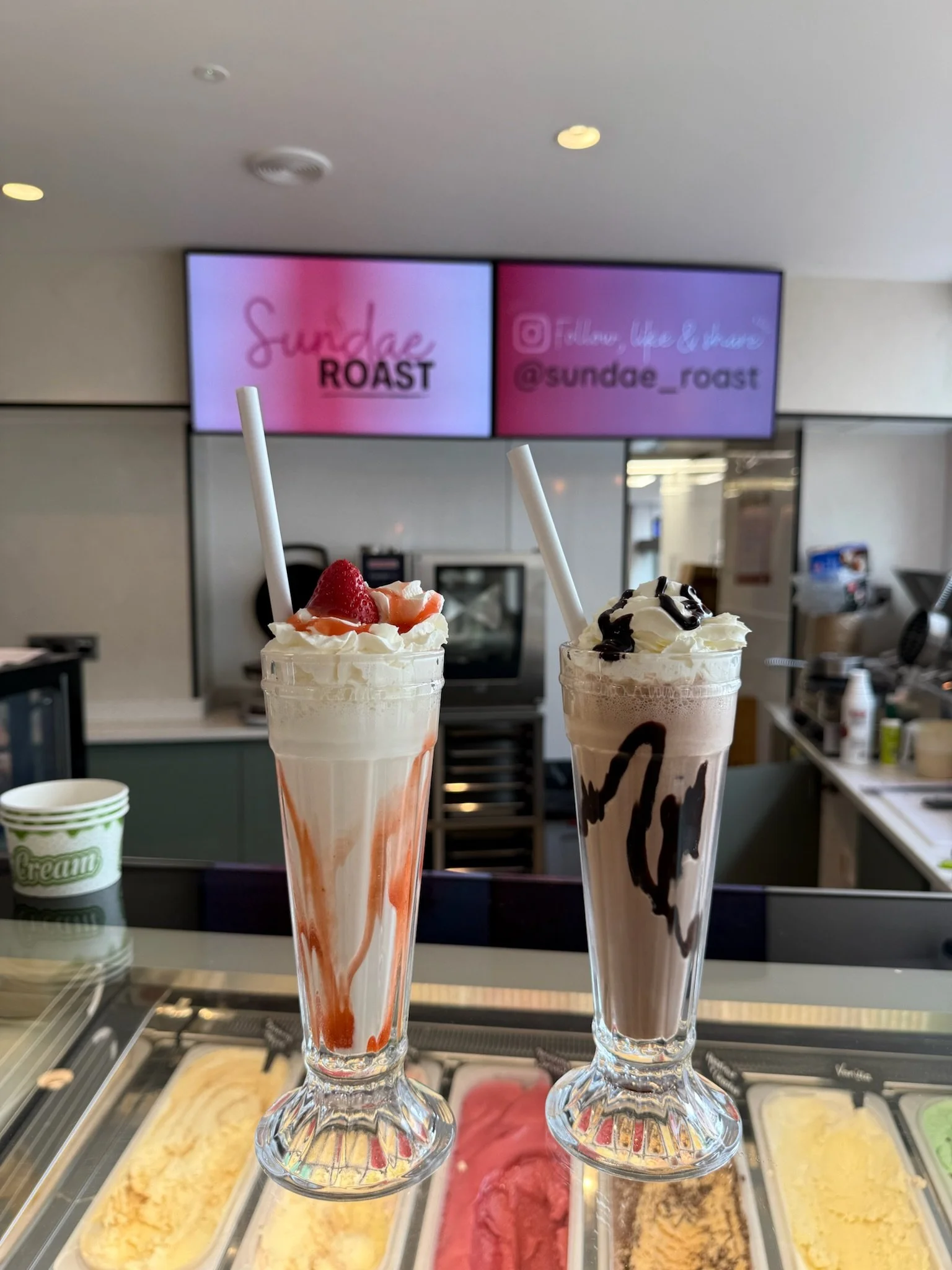 Two sundaes topped with whipped cream, strawberries, and chocolate syrup, with ice cream flavors in the background and a sign advertising Sundae Roast.