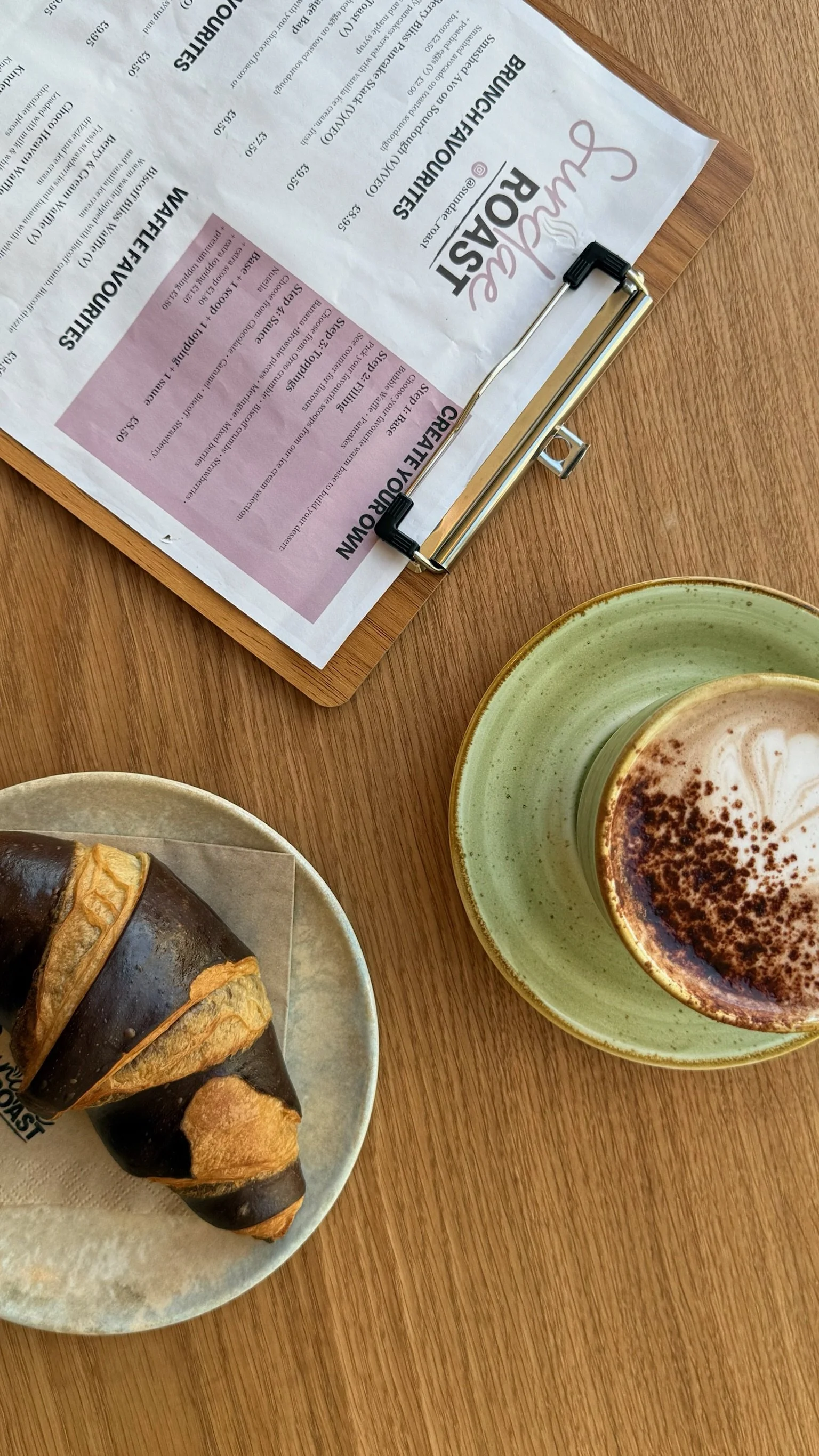 A wooden table with a menu on a clipboard, a cup of coffee with foam and sprinkled cocoa powder, and a pastry with chocolate glaze and almond slices.