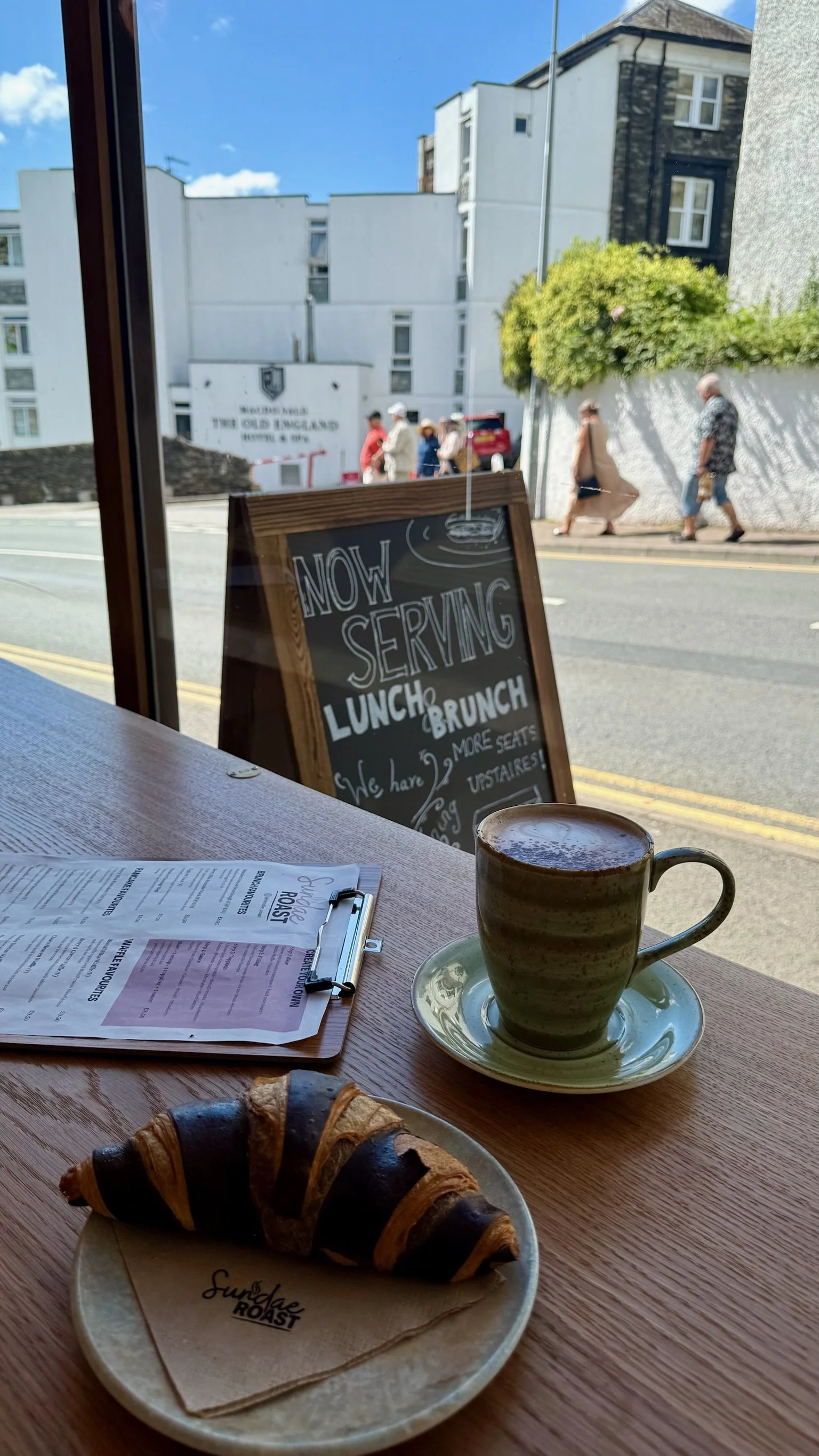 View from inside a cafe showing a croissant on a plate, a coffee cup on a saucer, and a menu on a clipboard on a wooden table. Outside the window, people walk on the sidewalk past a signboard that indicates the cafe is serving lunch and brunch. The s