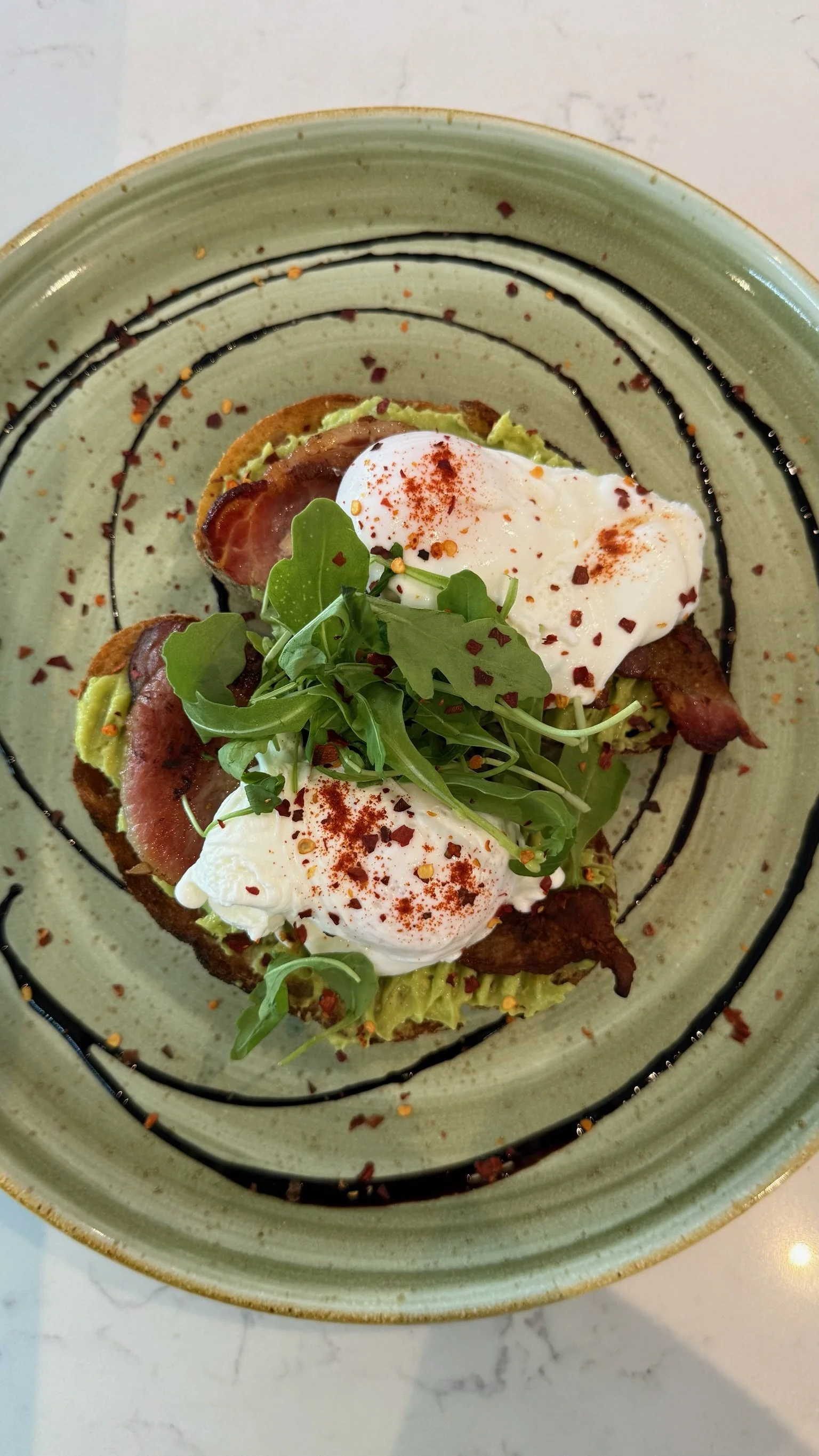 Open-faced sandwich topped with poached eggs, arugula, bacon, and avocado on a green plate with black sauce drizzle.