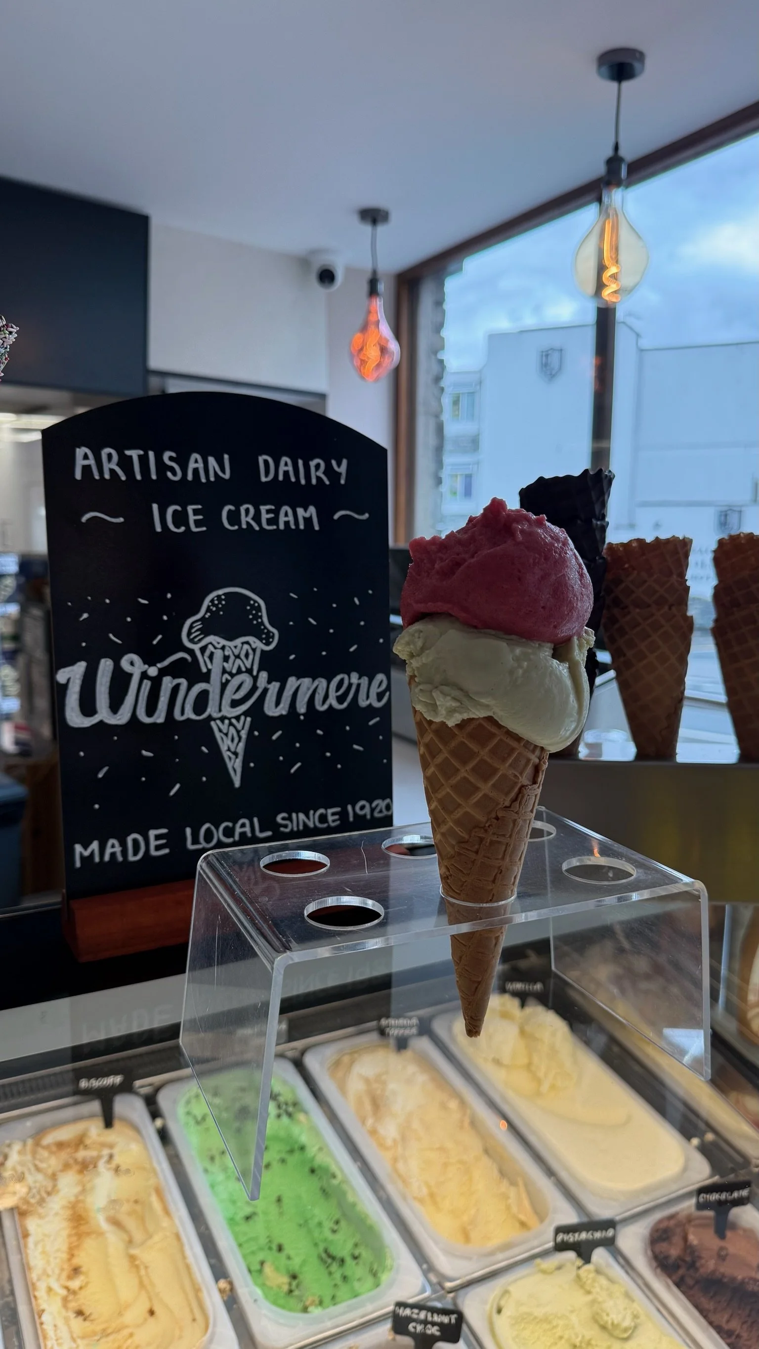 A display of various flavors of ice cream in a shop, with a sign that reads "Artisan Dairy Ice Cream" and a cone with three scoops of ice cream in front of the sign.