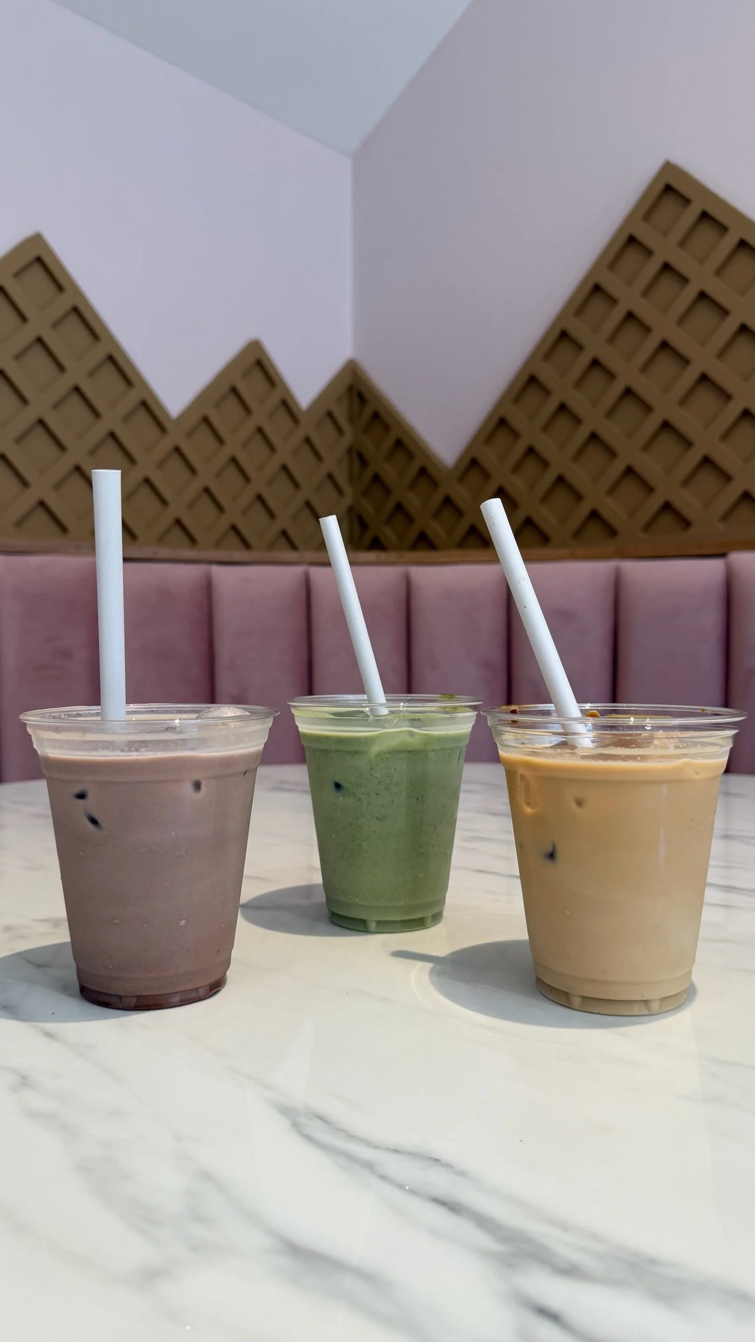Three cups of iced matcha latte, chocolate, and coffee drinks on a white marble table in a cafe with a pink cushioned seating and decorative wooden wall panels in the background.