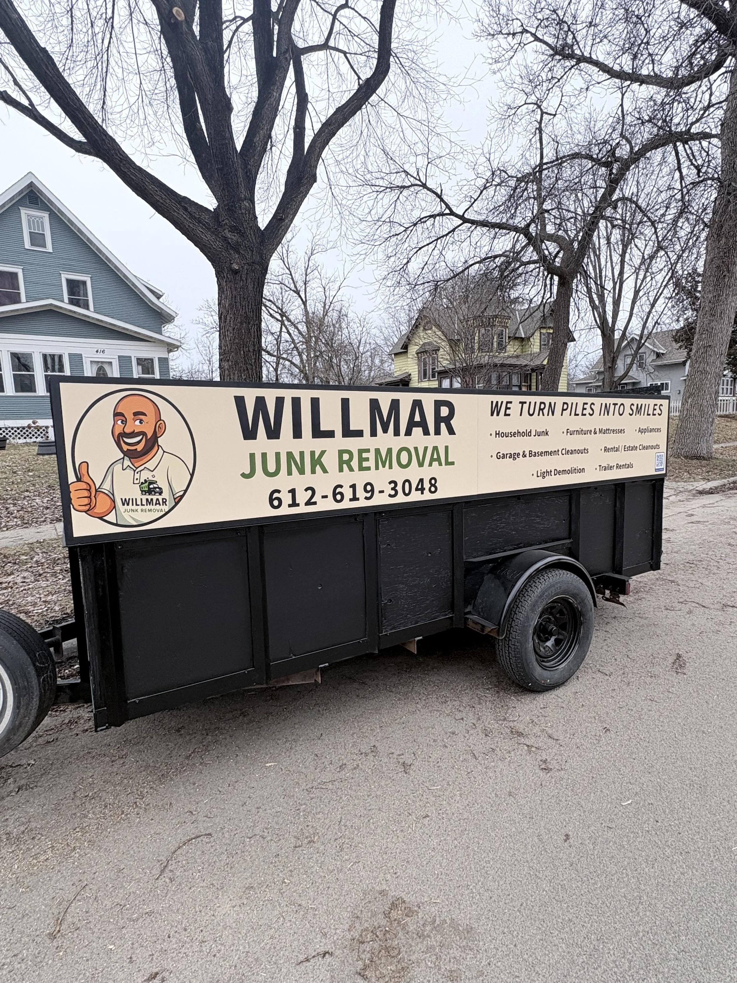 Black trailer with a sign advertising Willmar Junk Removal, featuring a cartoon man giving a thumbs up, along with phone number and list of services like household junk, furniture, appliances, garage, basement, rental cleanouts, light demolition, trailer rentals.