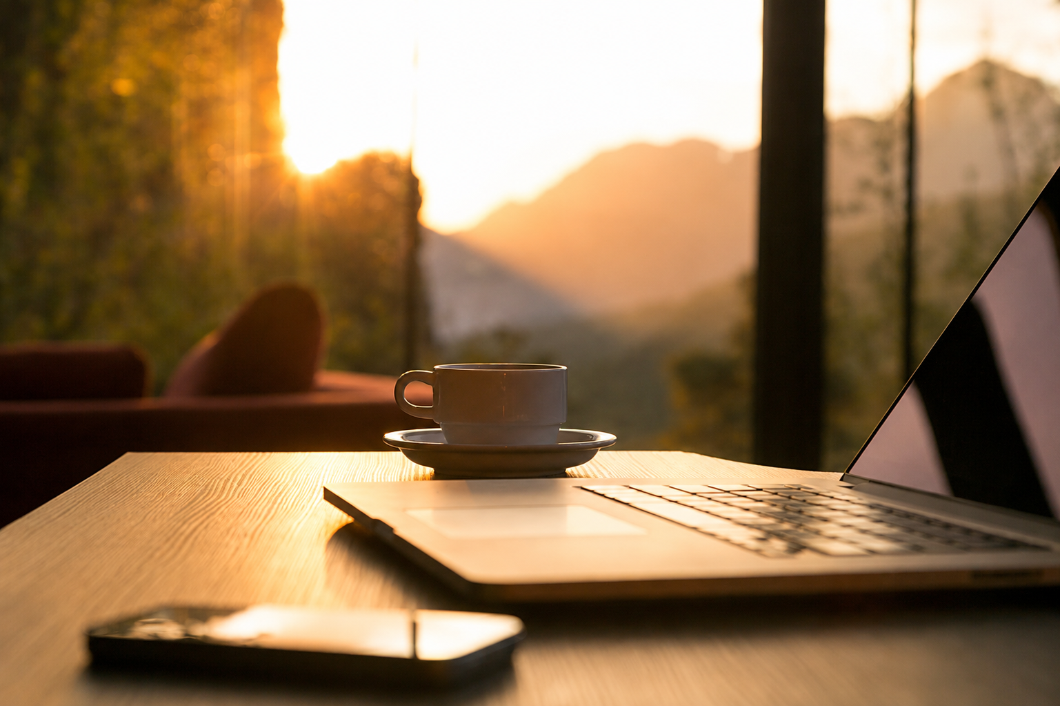 A laptop, a coffee cup on a saucer, and a smartphone on a wooden table in front of a large window with a view of trees and mountains at sunset.