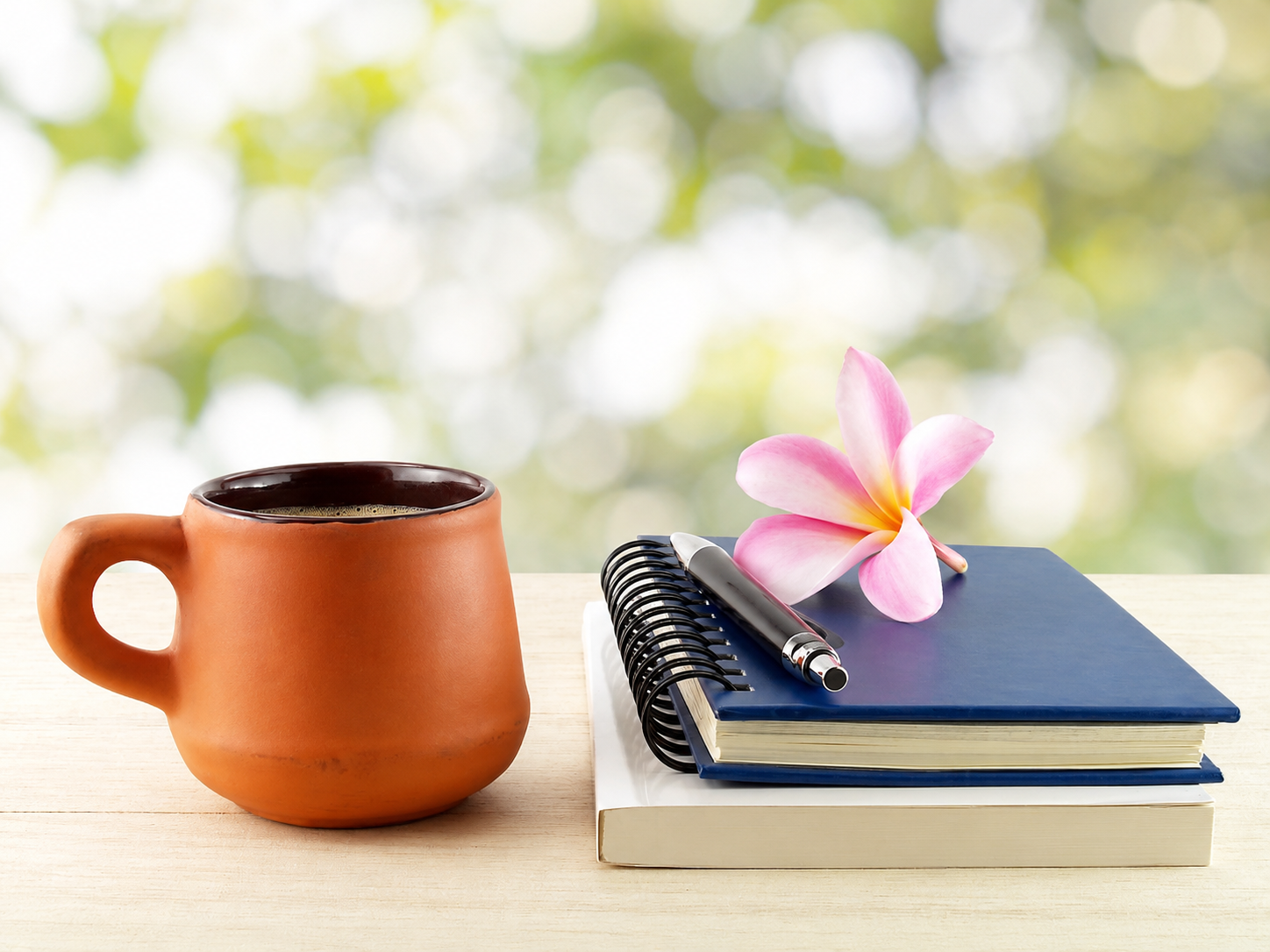 A terracotta mug filled with black coffee, a blue notebook, a black and silver pen, a pink flower, and a white and yellow book on a light wooden table against a blurred green outdoor background.