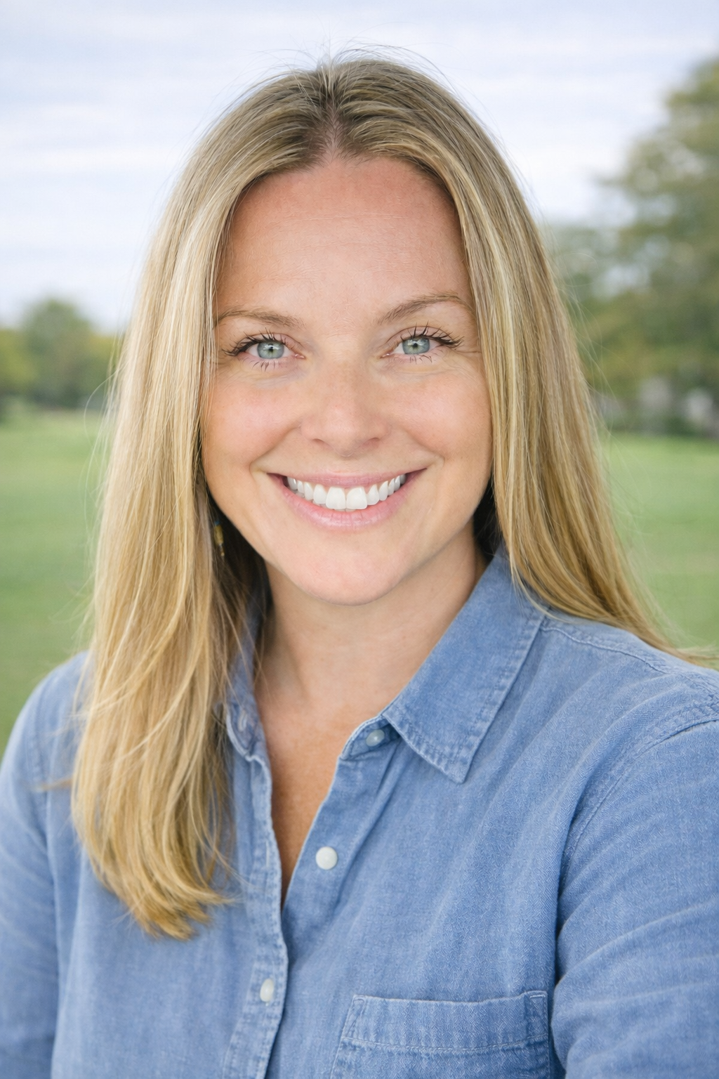 A woman with long blonde hair and blue eyes smiling outdoors, wearing a blue denim shirt, with a blurred green park background.