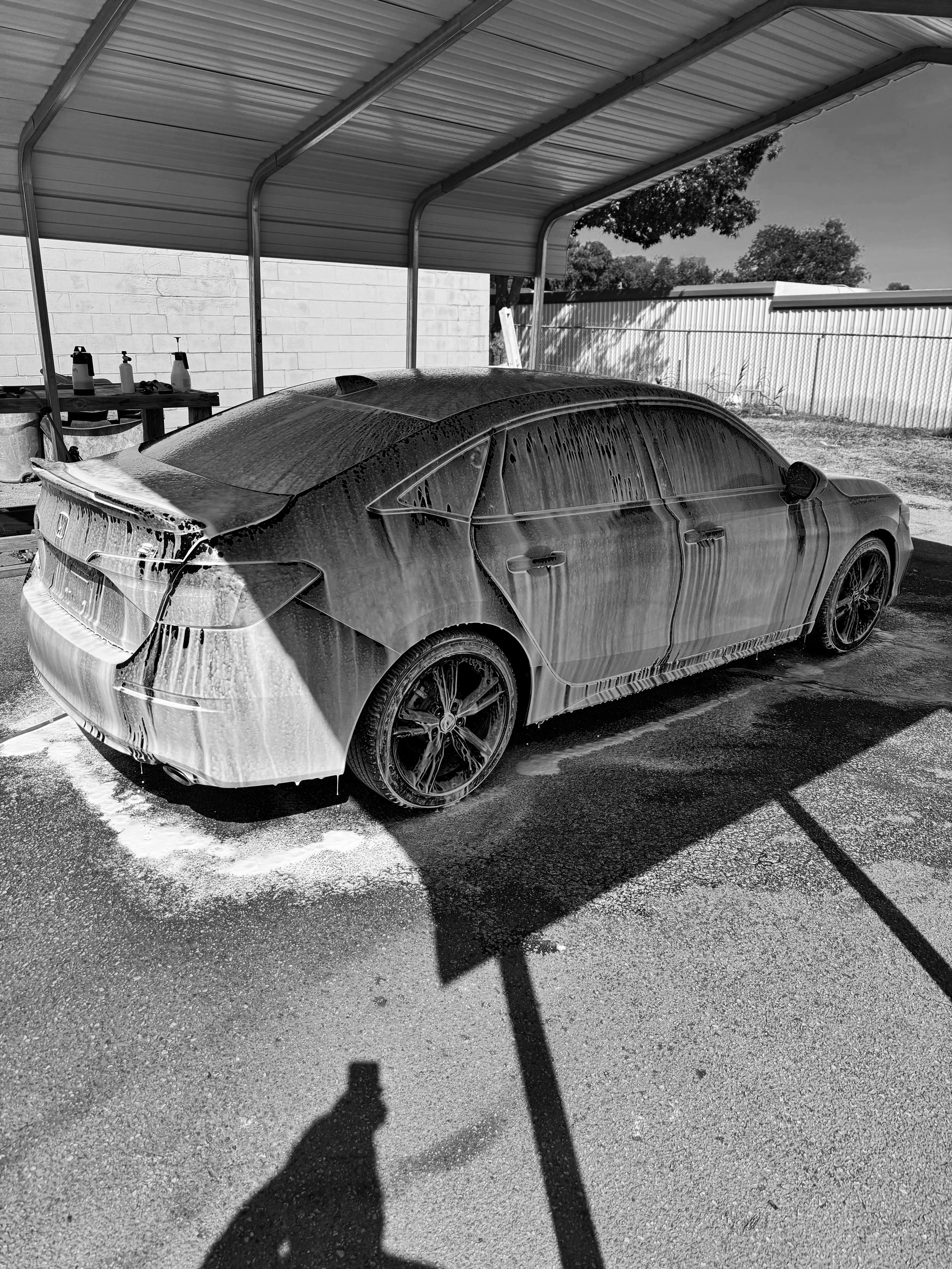 A black car covered in soap suds under a carport, with soap dripping and foam on the ground.