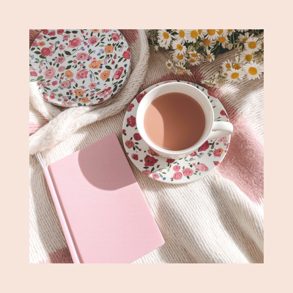 A floral teacup and saucer filled with tea, a pink notebook, a plate with a floral pattern, and a bunch of daisies arranged on a cozy blanket.