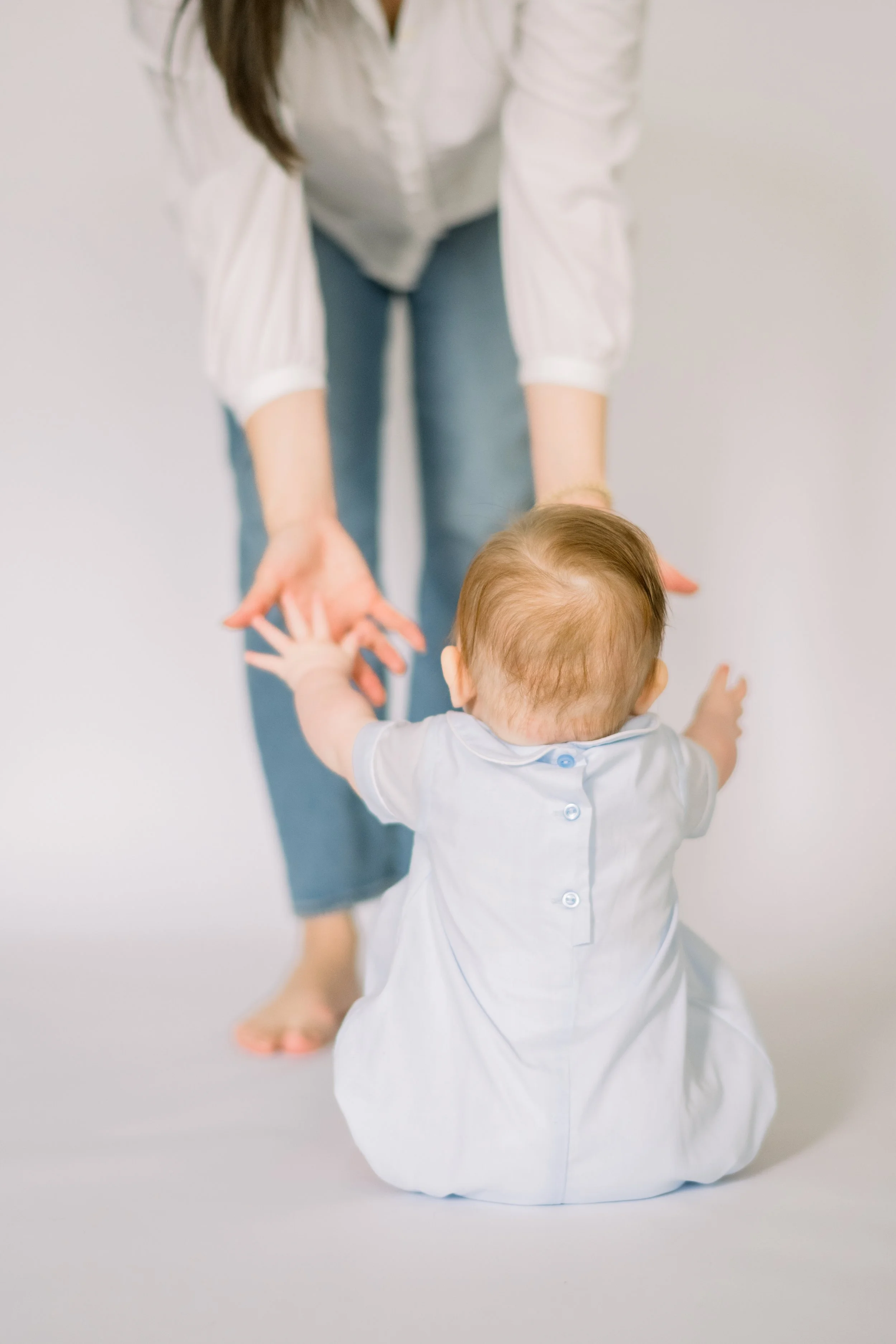 A young child kneeling on the floor with arms outstretched towards a woman, who is bent over reaching down to hold the child's hands. The woman is wearing a white shirt and blue jeans, and the child is dressed in a white shirt. The background is plain and light-colored.
