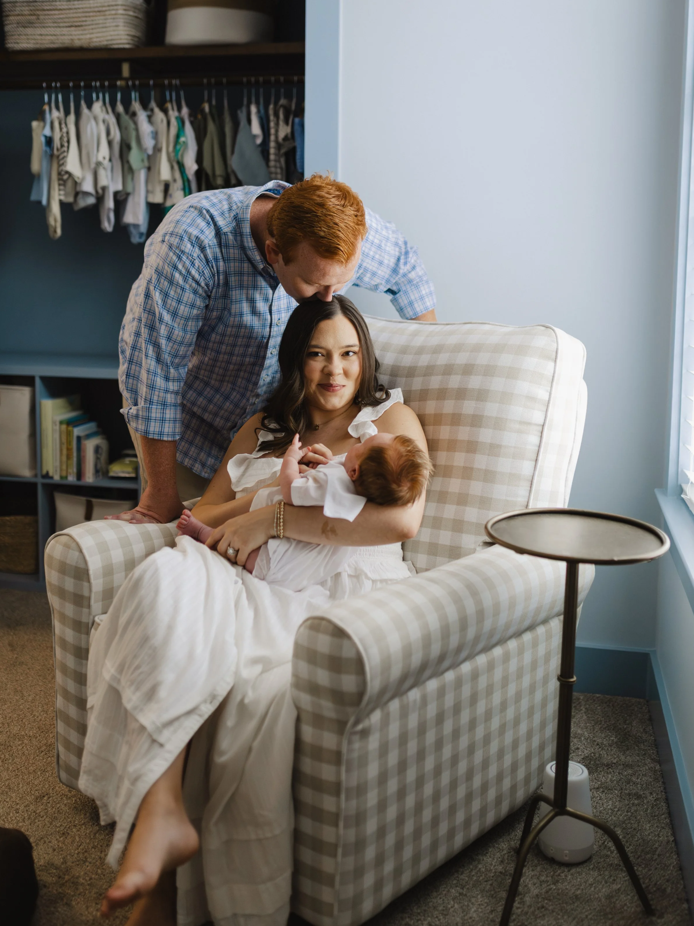 A woman is lying on a beige and white checkered armchair, breastfeeding a baby. A man leans over her, kissing her forehead. There is a small round side table with a diffuser beside the armchair, and a bookcase with books and baskets in the background. An open closet with hanging clothes is visible behind them.