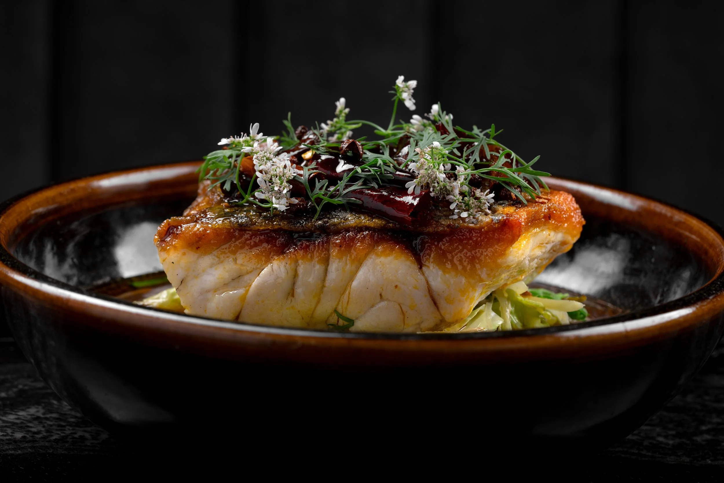 Pan-seared fish with crispy skin, dark sauce, and fresh herb flowers, served in a ceramic bowl at Animae San Diego