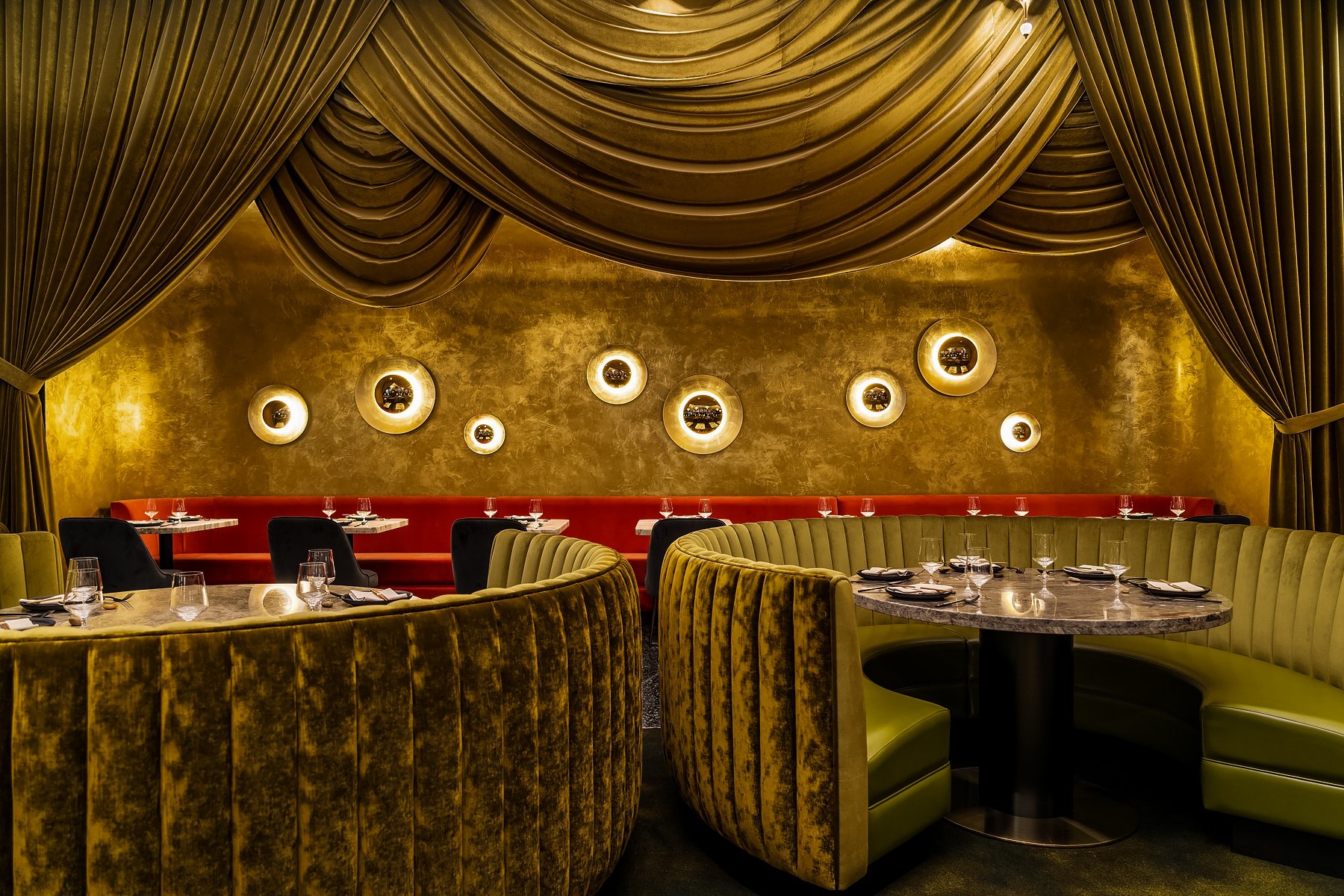 Animae dining room featuring draped gold velvet walls, circular porthole sconces, curved velvet booths, and intimate table settings