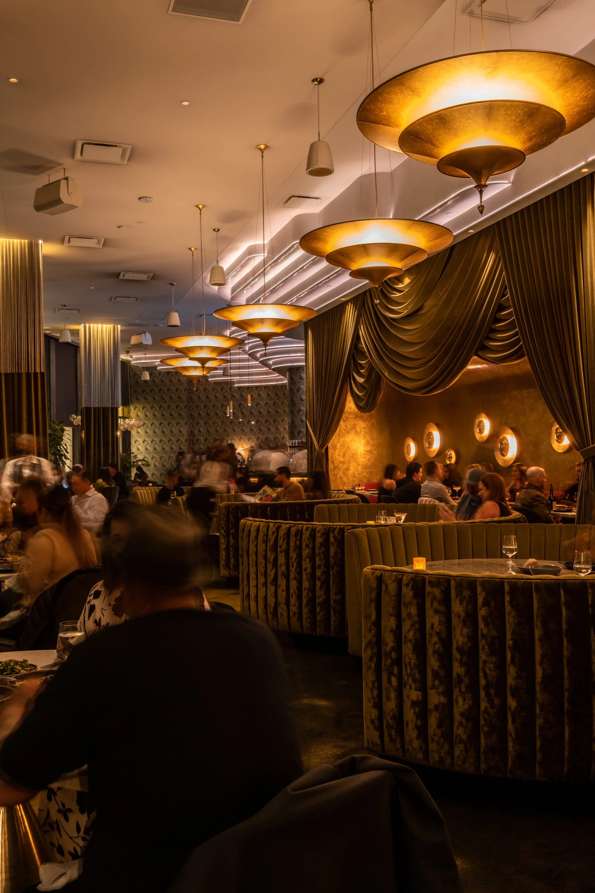 Animae dining room at full capacity during evening service, with stacked gold pendant lights, draped olive velvet walls, circular booths, and a lively atmosphere, downtown San Diego