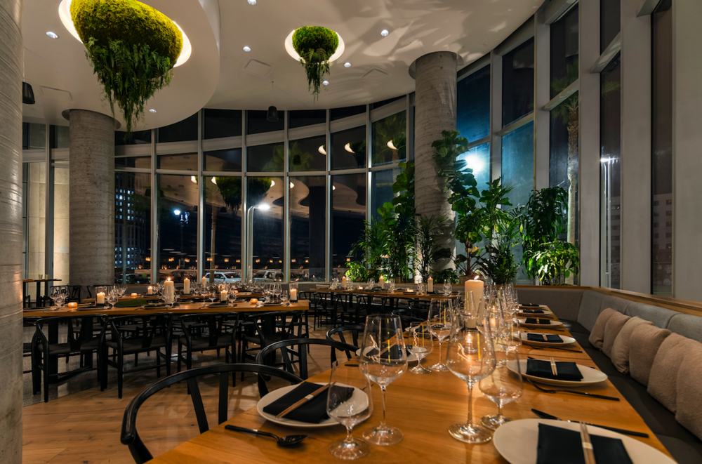 Nima private event space at Animae San Diego set for a group dinner at night, with tropical plants, floor-to-ceiling windows, and candlelit long tables