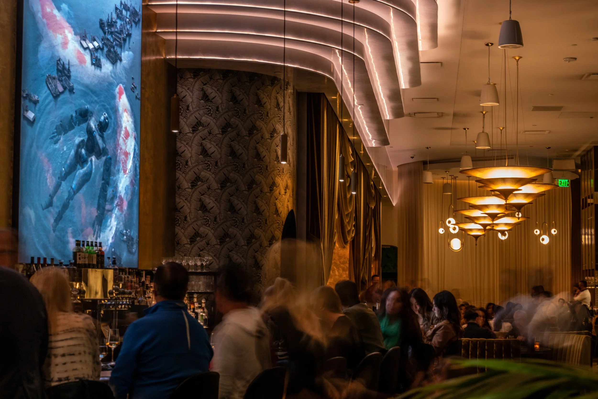 The dining room at Animae San Diego, featuring dramatic gold pendant lighting, Art Deco design, a large koi mural, and a full house of guests at dinner service