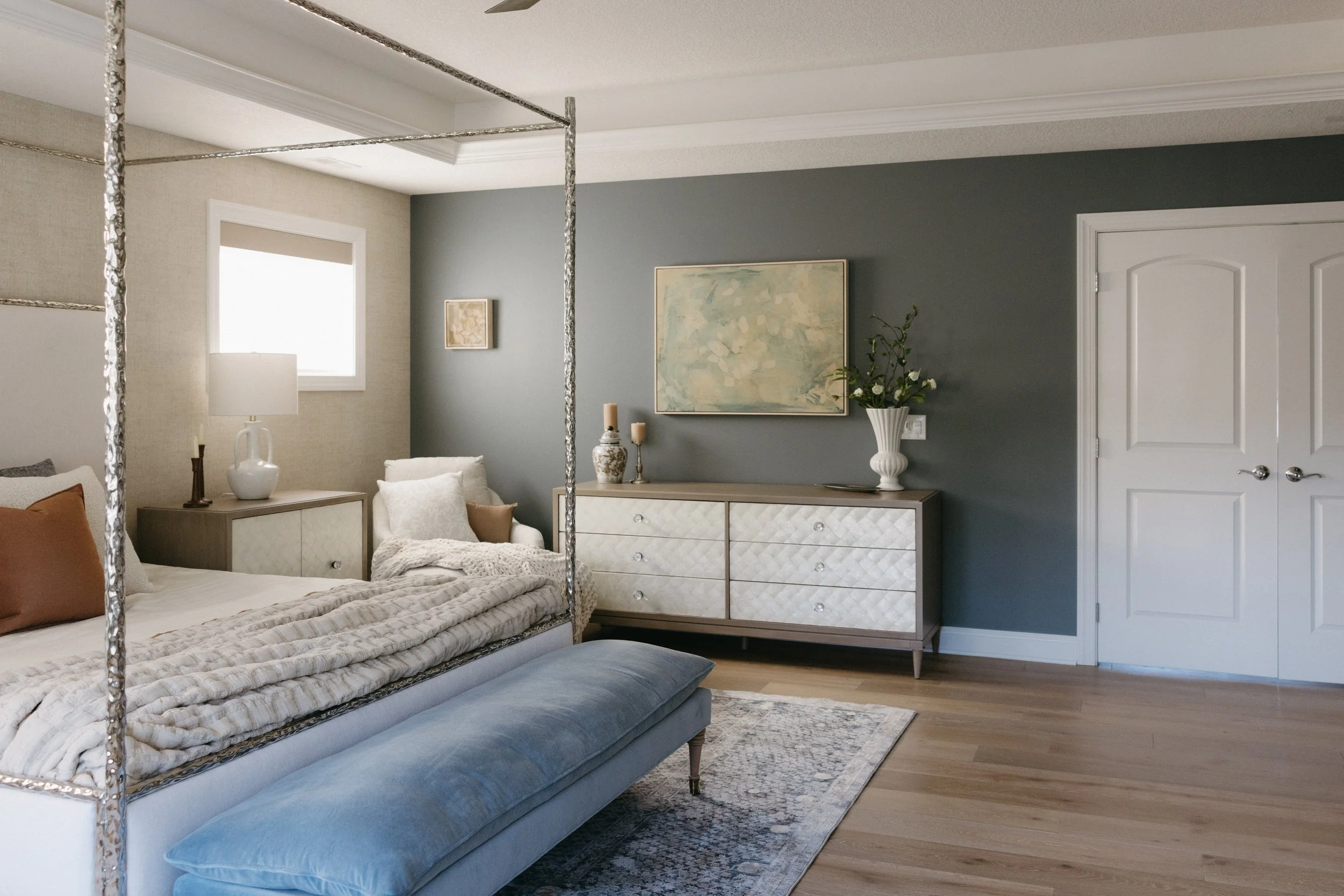 Primary bedroom designed by Texturious Designs featuring layered neutral furnishings, blue accents, custom furniture, and warm wood flooring.