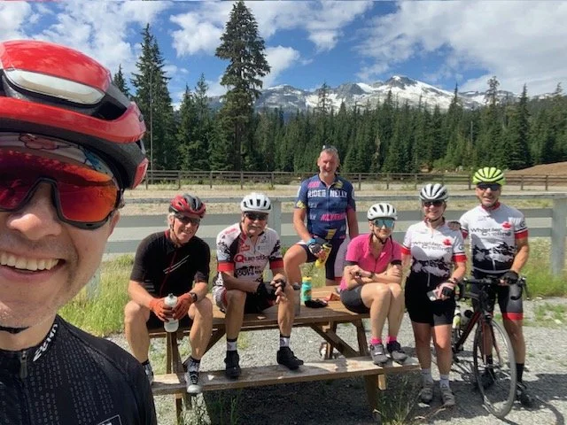 Group of six WCC cyclists taking a break at the top of the Callaghan Valley, with snow-capped mountains and pine trees in the background.
