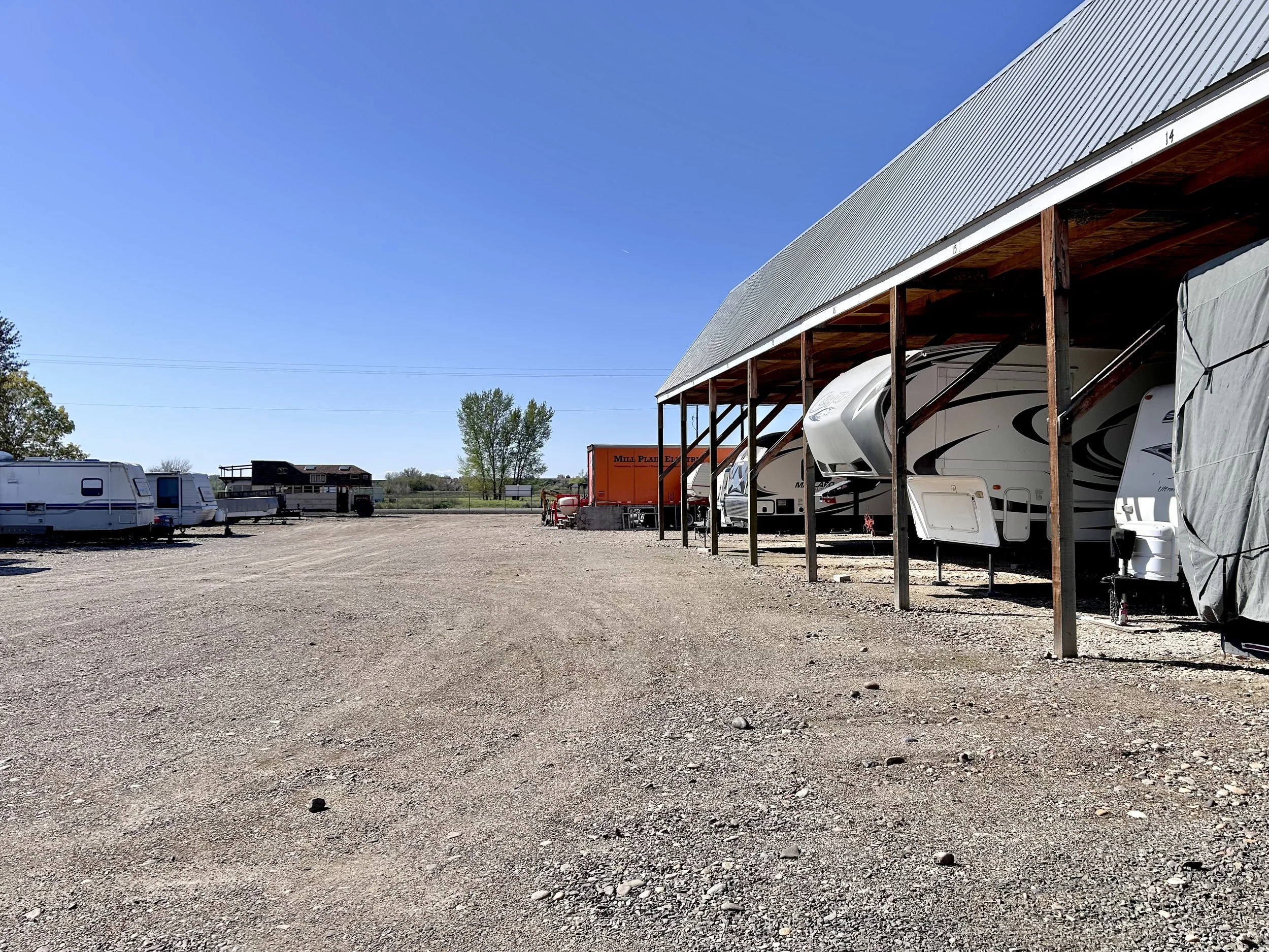A gravel lot with several parked travel trailers under a carport, some construction equipment, trees, and a clear blue sky.
