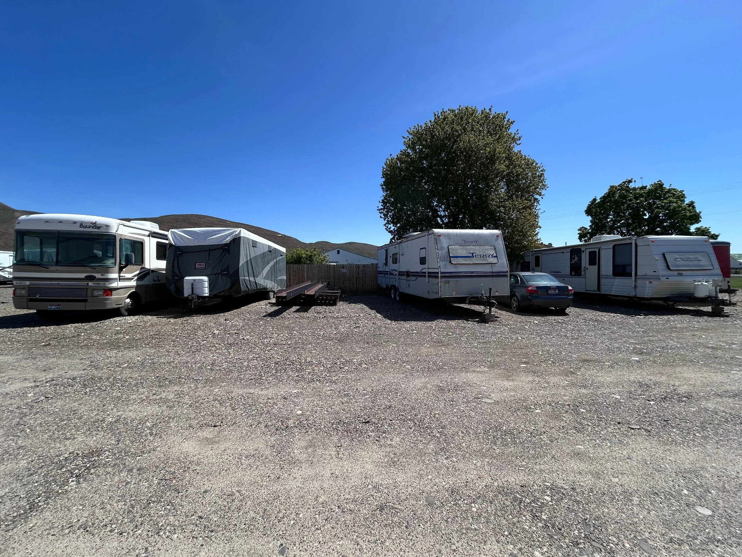 A parking lot with several recreational vehicles and trailers parked on a gravel surface under a clear blue sky. There are a few trees and a hillside in the background.