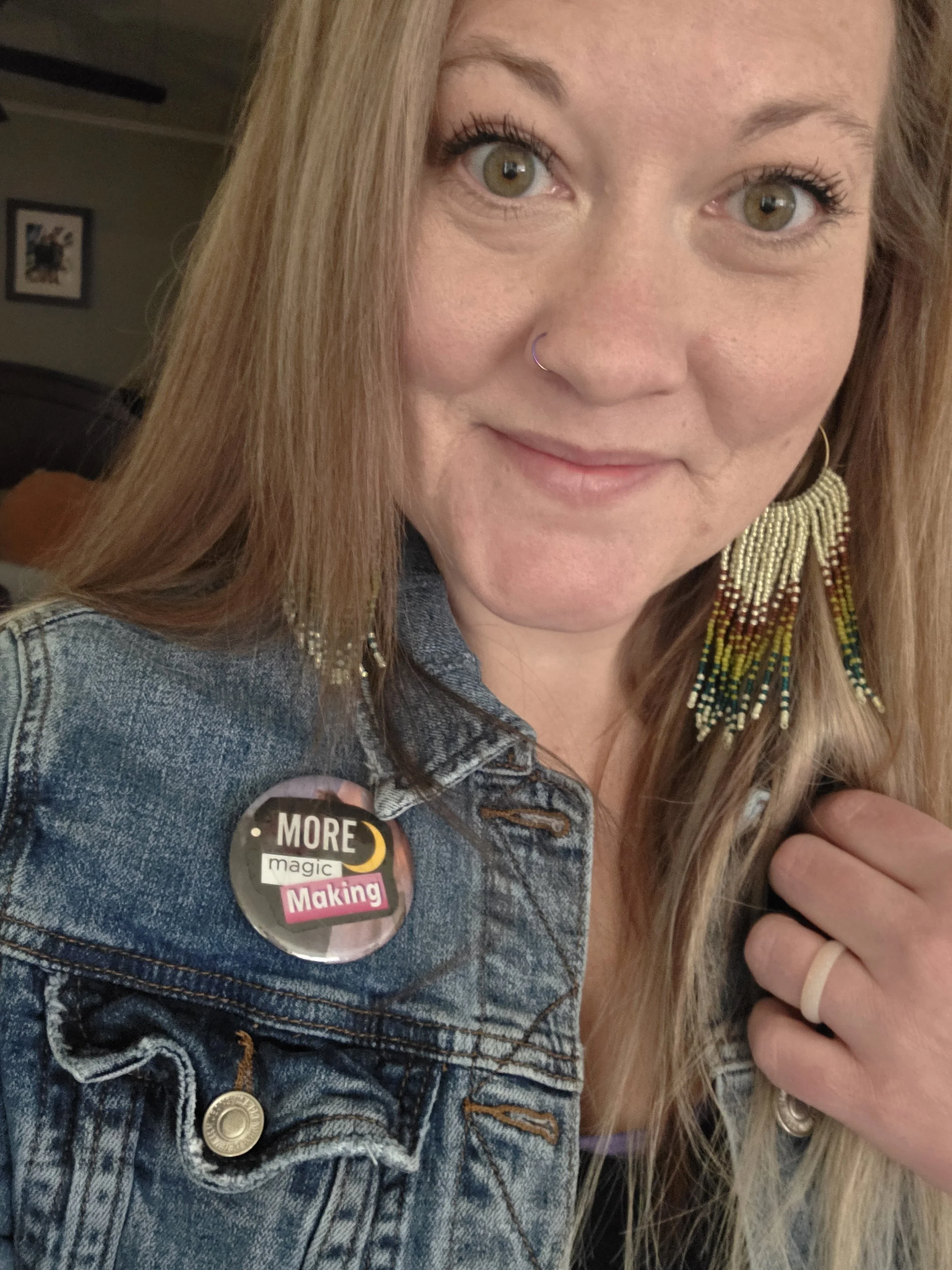 A woman with light brown hair and green eyes wearing a denim jacket with a button that says 'MORE magic Making,' beaded earrings, a ring, and a nose ring, smiling at the camera.