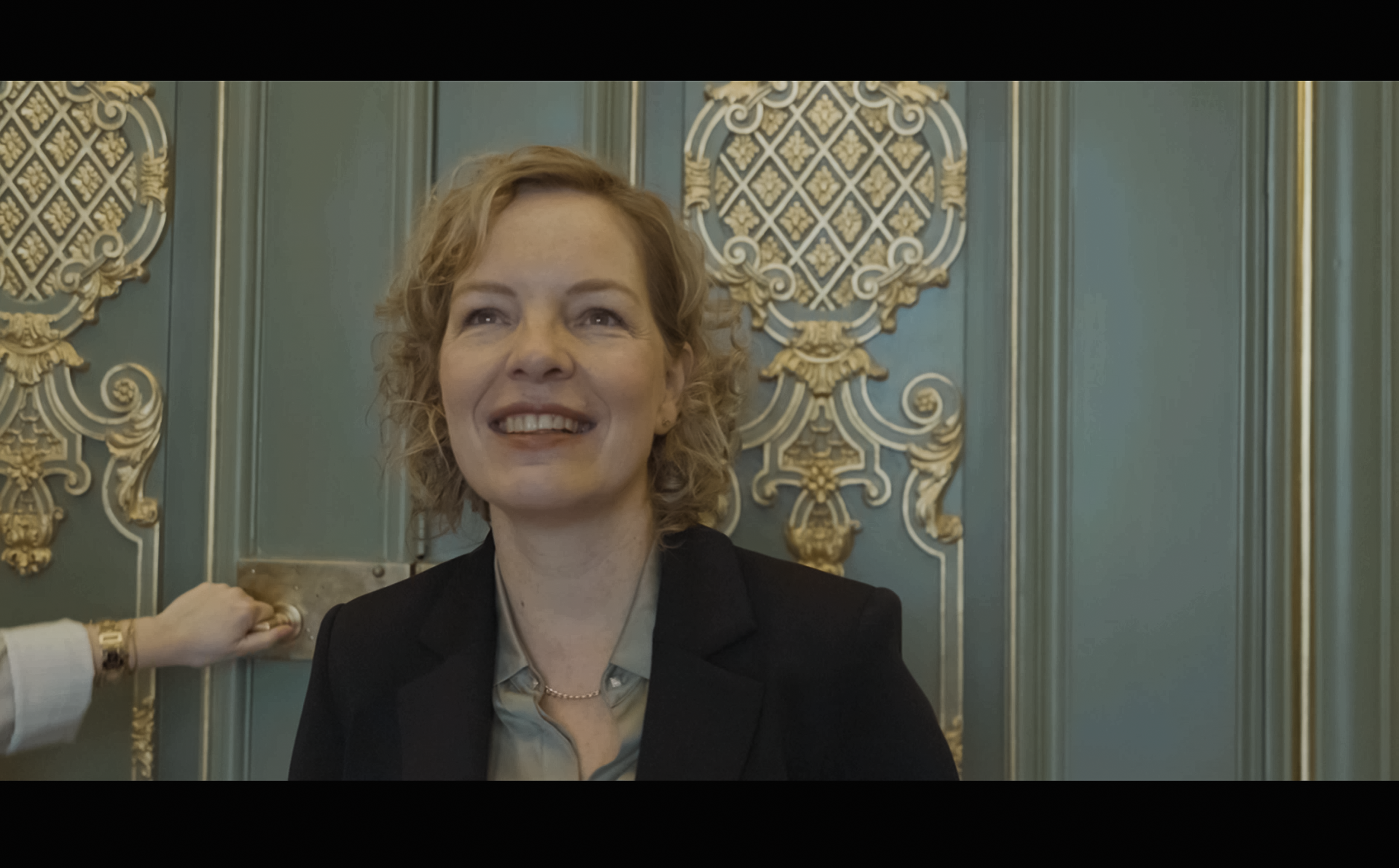 Een vrouw met korte, krullende blonde haren glimlacht en kijkt omhoog, terwijl iemand haar hand vasthoudt. Ze zit voor een decoratieve blauwe muur met goudkleurige versieringen. Beatrice de Graaf