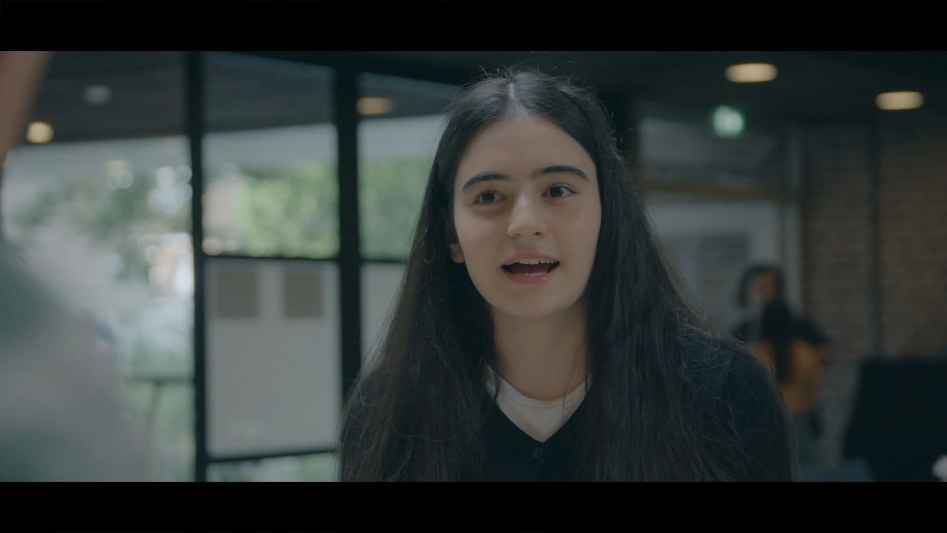 Een jonge vrouw met lang, donker haar spreekt in een gesprek, sitting in een ruimte met grote ramen en achtergrond met een persoon die op een stoel zit.