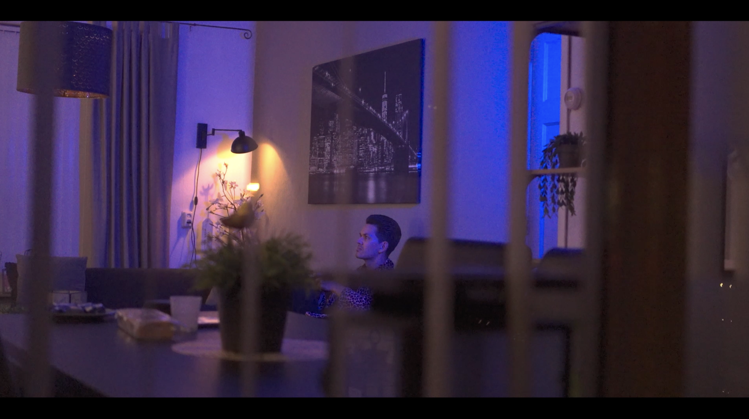 Een man zit aan een eettafel in een sfeervol verlichte kamer met blauwe en gele verlichting. Op de achtergrond hangt een grote foto van een brug bij nacht, en er is een lamp aan de muur. De tafel heeft een plant en andere items.