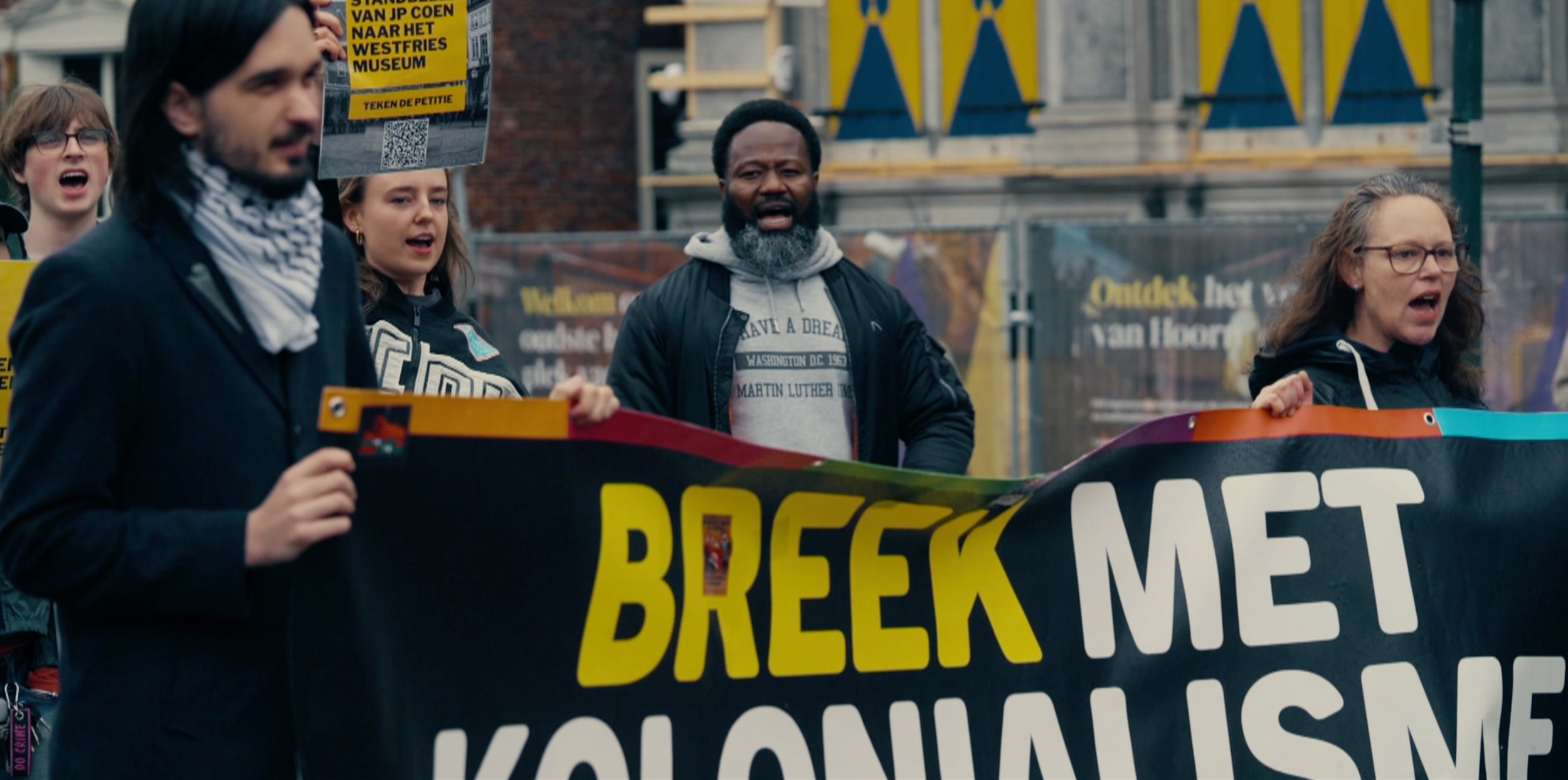 Mensen demonstreren buiten tijdens een protest. Ze houden een grote banner met de tekst 'BREAK MET' en andere woorden die niet volledig zichtbaar zijn. Sommigen roepen of zingen, en de sfeer is betrokken en emotioneel.