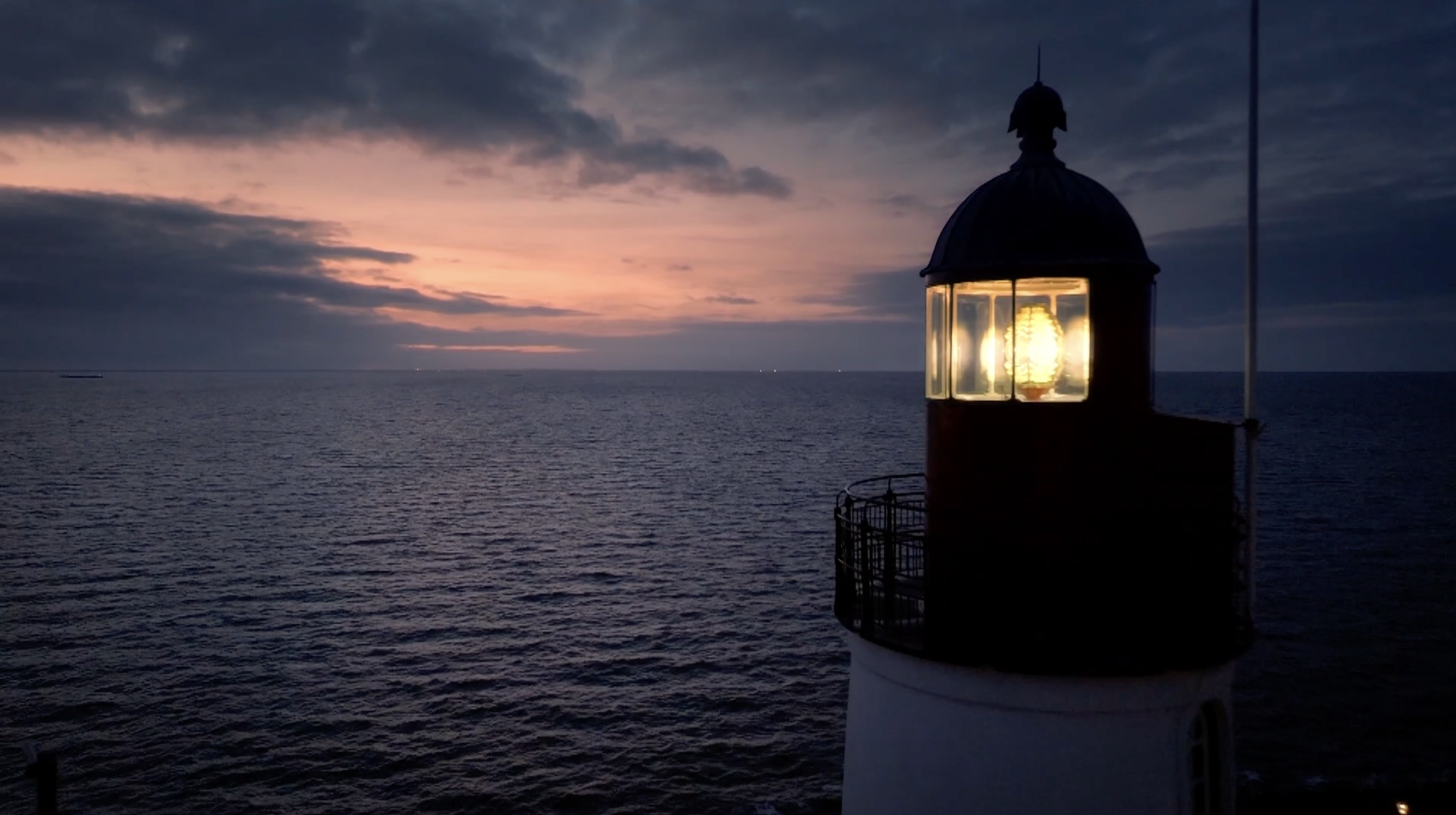 Een vuurtoren aan de rand van de oceaan tijdens zonsondergang, met een licht dat helder schijnt door de ramen van de vuurtoren, donkere wolken in de lucht.