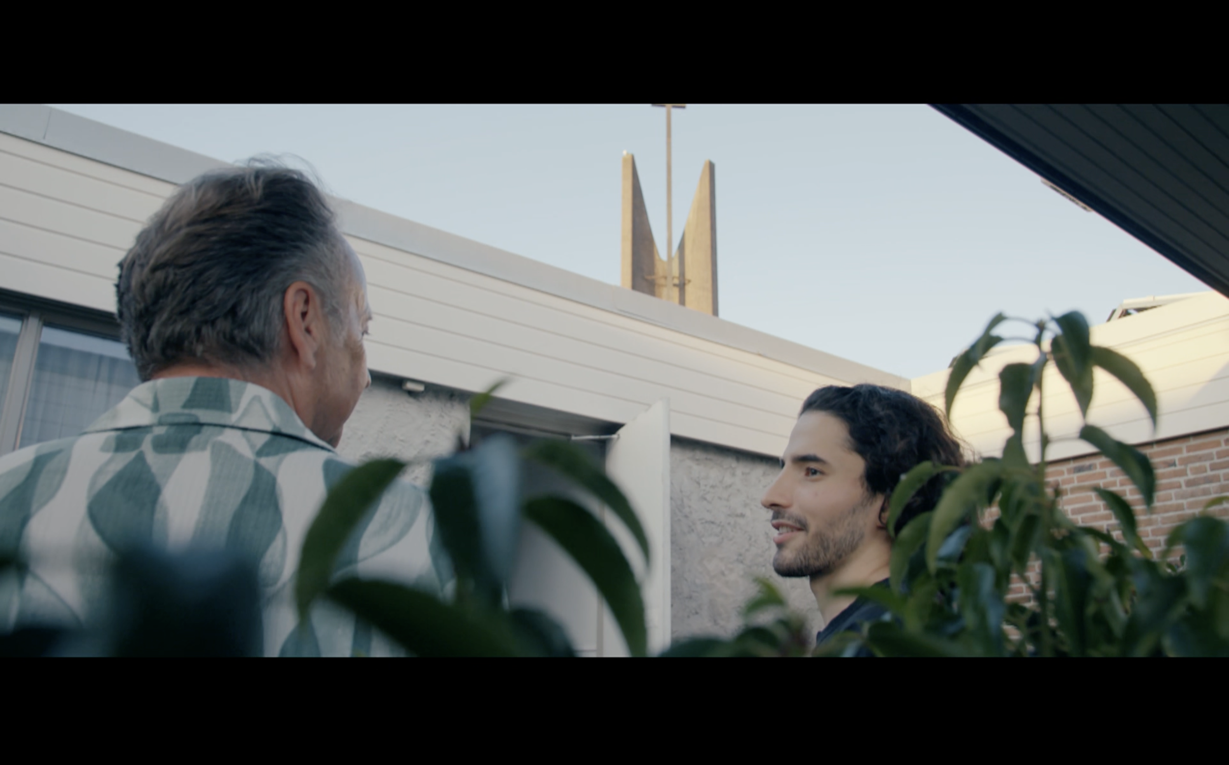 Twee mannen praten op een terras met planten voor hen, met woongebouwen en een kerktoren op de achtergrond.