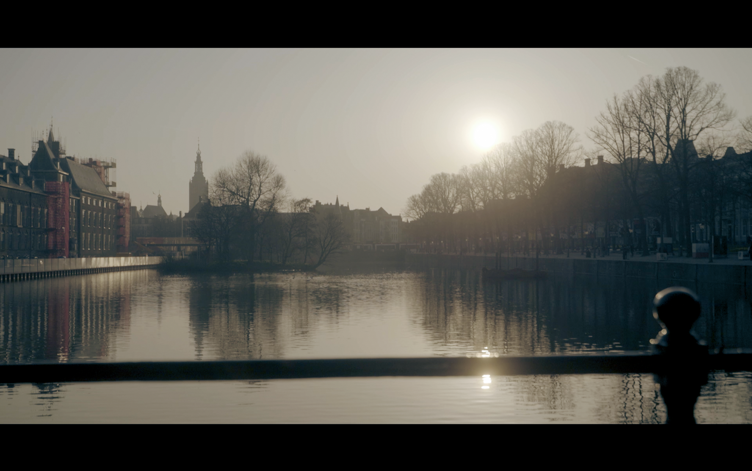 Een kalme rivier met gebouwen en bomen langs de oever, de zon die opkomt of ondergaat en de silhouetten van de gebouwen en bomen tegen de lucht.