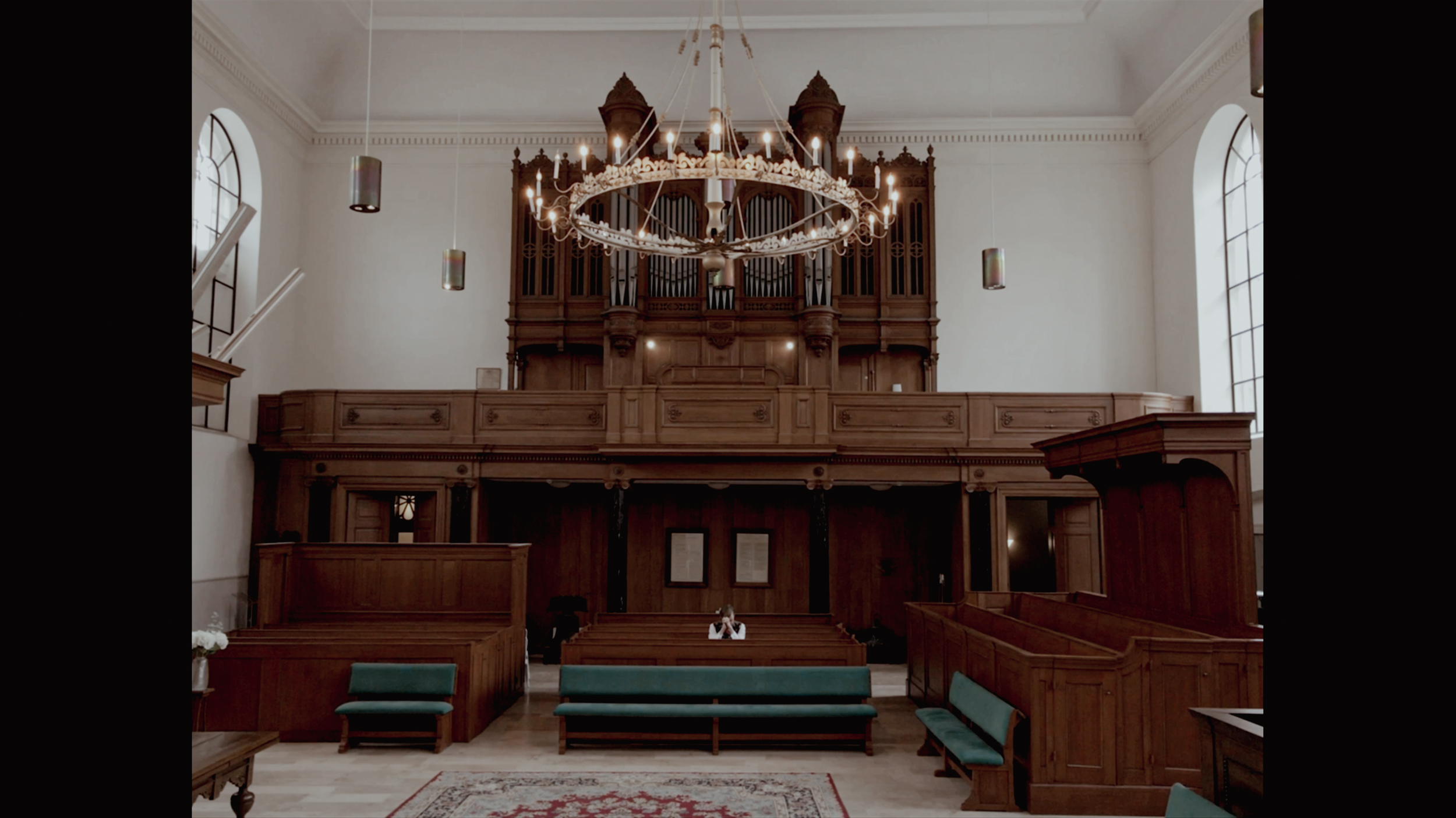 Interieur van een kerk met grote houten orgel en chandeleer aan het plafond, met een vrouw zittend op een bank in de zaal.
