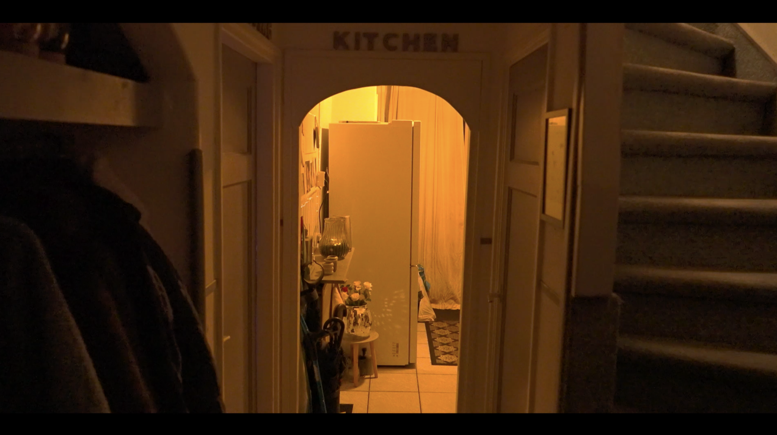 Een lichte gang met een deur die toegang geeft tot een woonkamer. De kamer is verlicht met een warme, geel-oranje gloed die een gezellige sfeer uitstraalt.