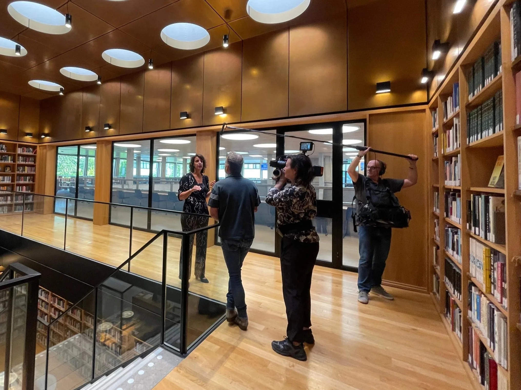 Mensen interviewen en filmen in een bibliotheek met houten boekenplanken, grote ramen en een houten plafond met ronde uitsparingen.