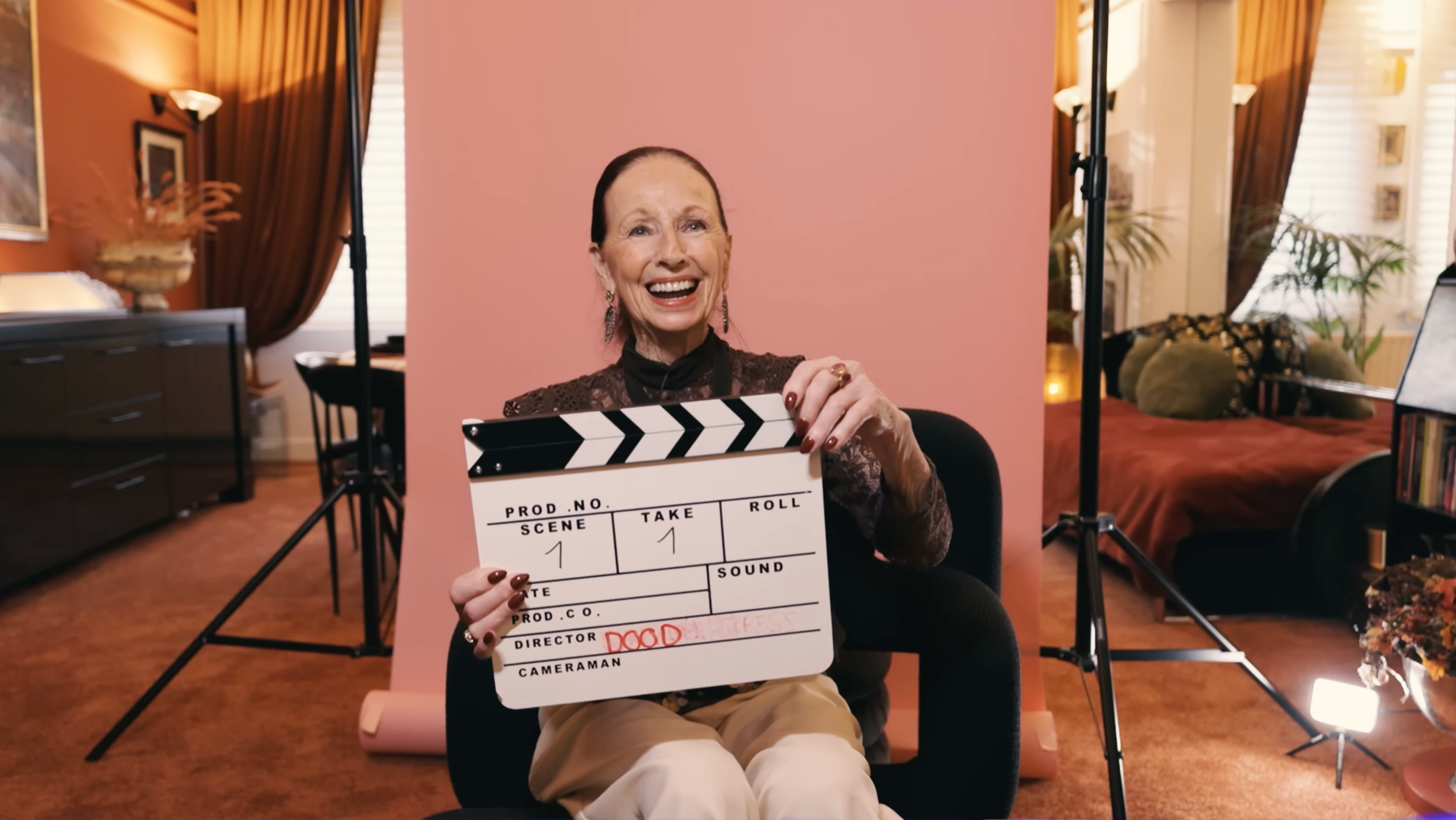 Vrouw die lachend een filmclapje vasthoudt, zittend in een sfeervolle kamer met rode en groene decoraties.
