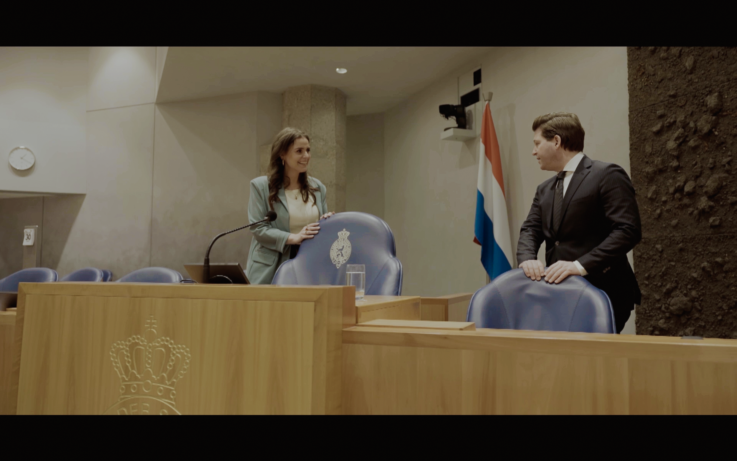 Een vrouw en een man staan in een vergaderruimte, glimlachend naar elkaar. De vrouw staat achter een stoel met een kerkelijke staflogo, terwijl de man naast haar staat, tegen de tafel leunend. Achter hen staat een Nederlandse vlag, en er is een muur met een rotsachtige structuur.