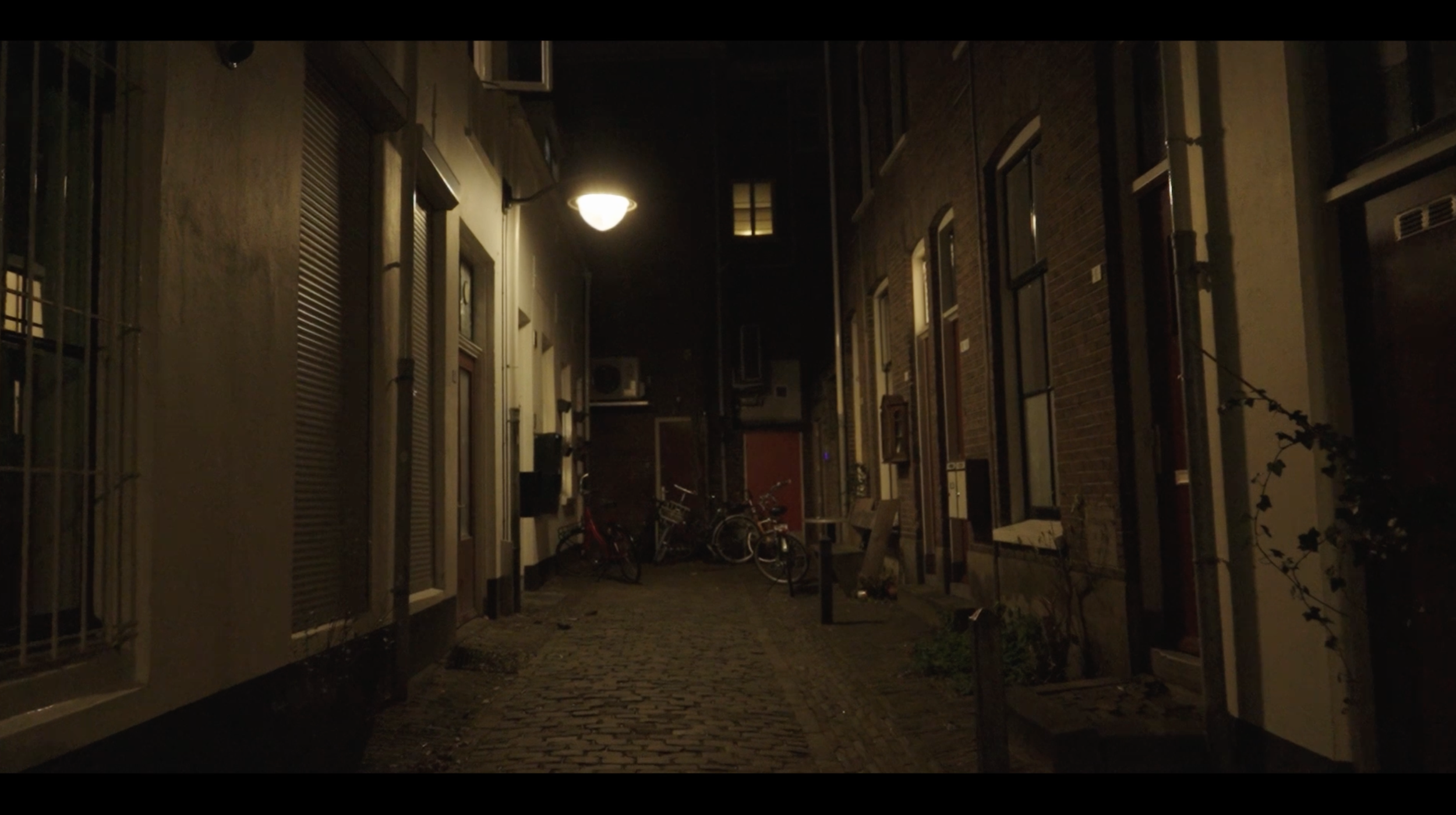 Een donker straatje met een straatlantaarn die licht werpt, omgeven door appartementen met ramen en fietsen.
