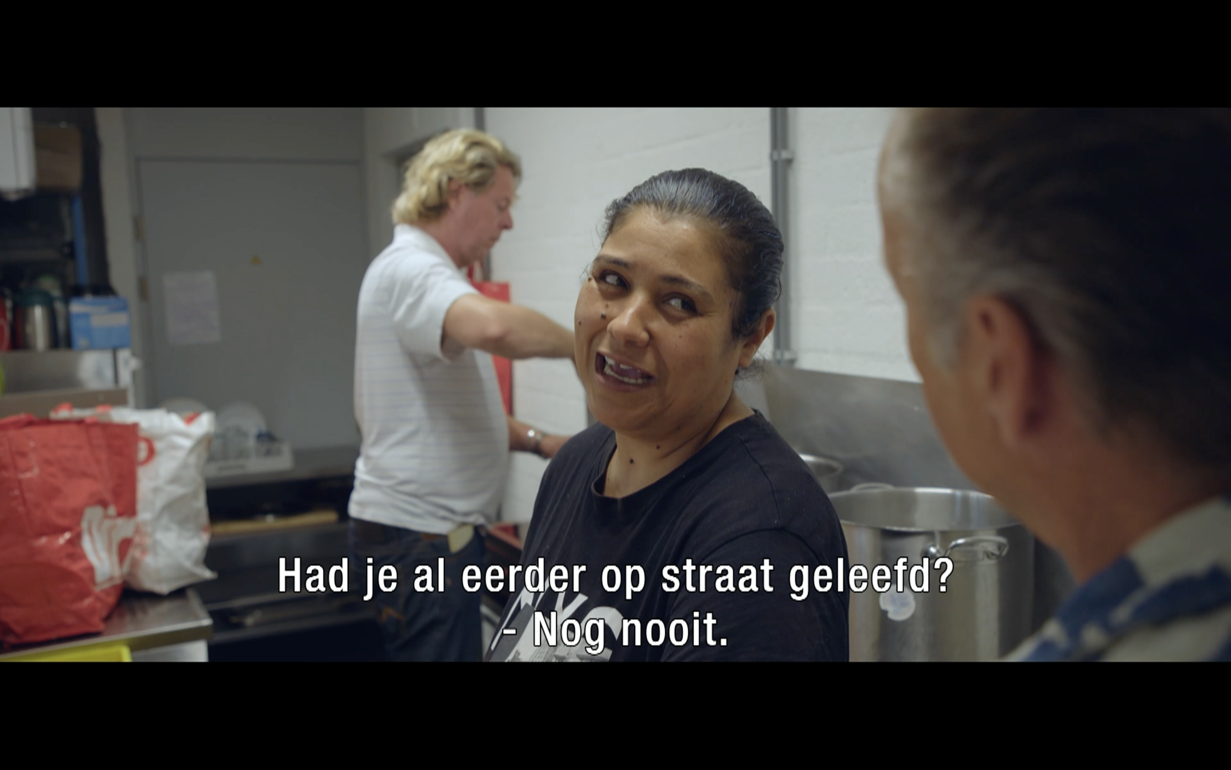 Drie vrouwen in een keuken, twee van hen praten, met ondertitels in het Nederlands. Eén vrouw kijkt verbaasd en lacht, de ander wijst naar haar. De achtergrond bevat keukenbenodigdheden en een witte muur.