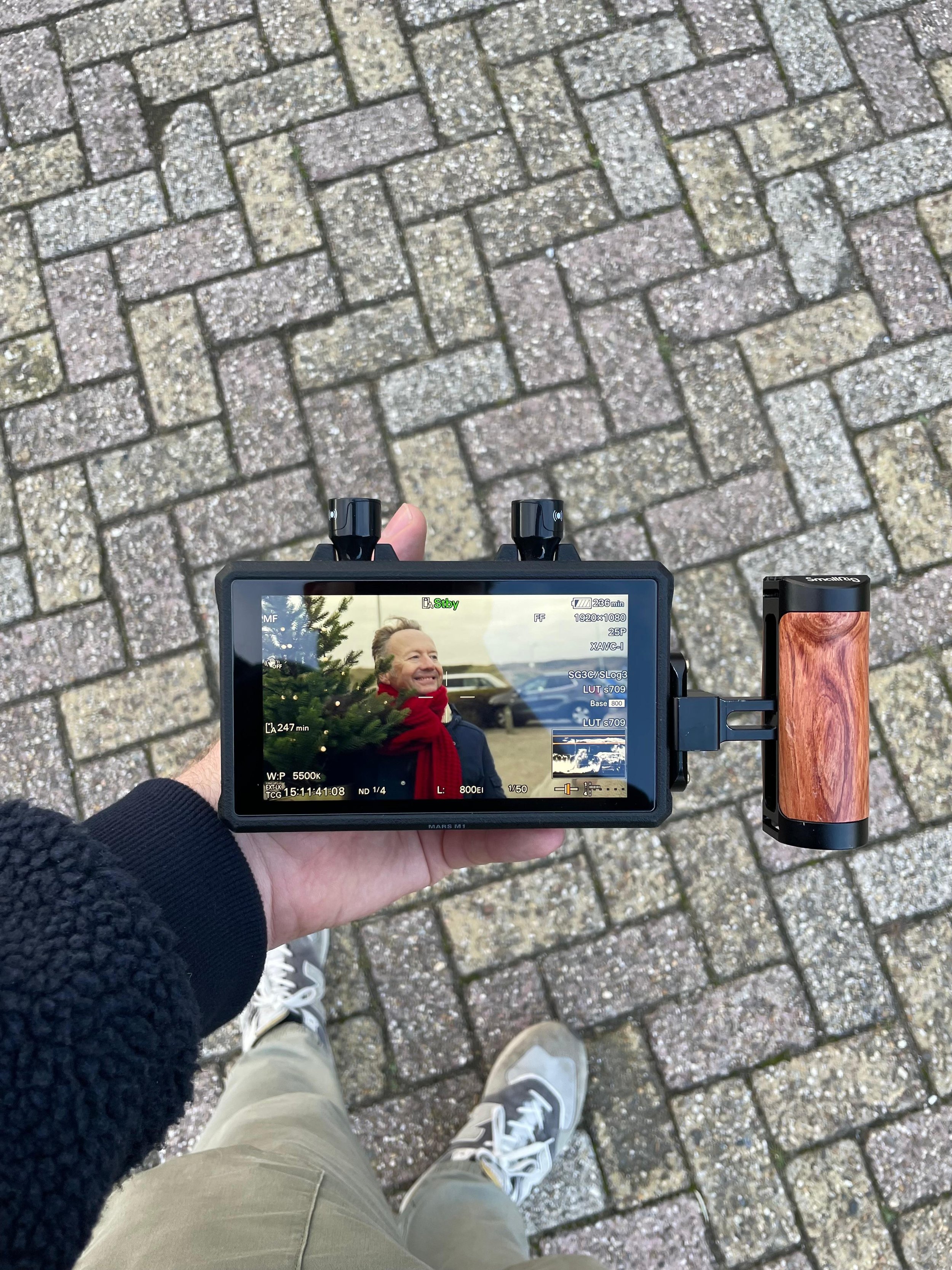 De foto toont een hand die een camera vasthoudt, gericht op een man die buiten staat met een kerstboom en een rood sjaal, terwijl hij glimlacht. De achtergrond bestaat uit een stenen bestrating.
