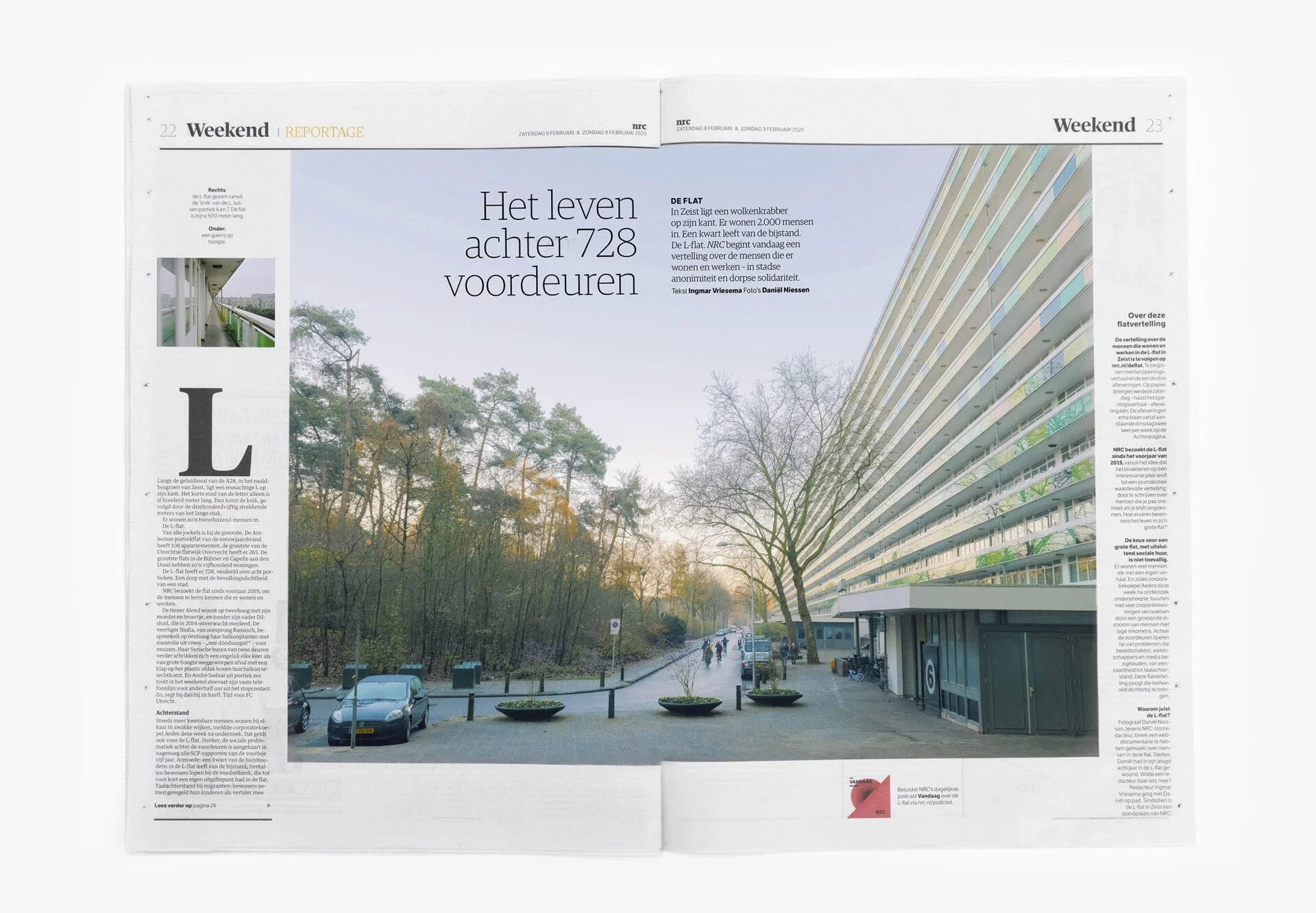 Een foto van een stedelijk landschap met een grote moderne appartementencomplex aan de rechterkant en een parkeerplaats met enkele auto's en een pad met wandelende mensen op de voorgrond. Bomen zonder bladeren zijn zichtbaar, en de lucht is lichtblauw.