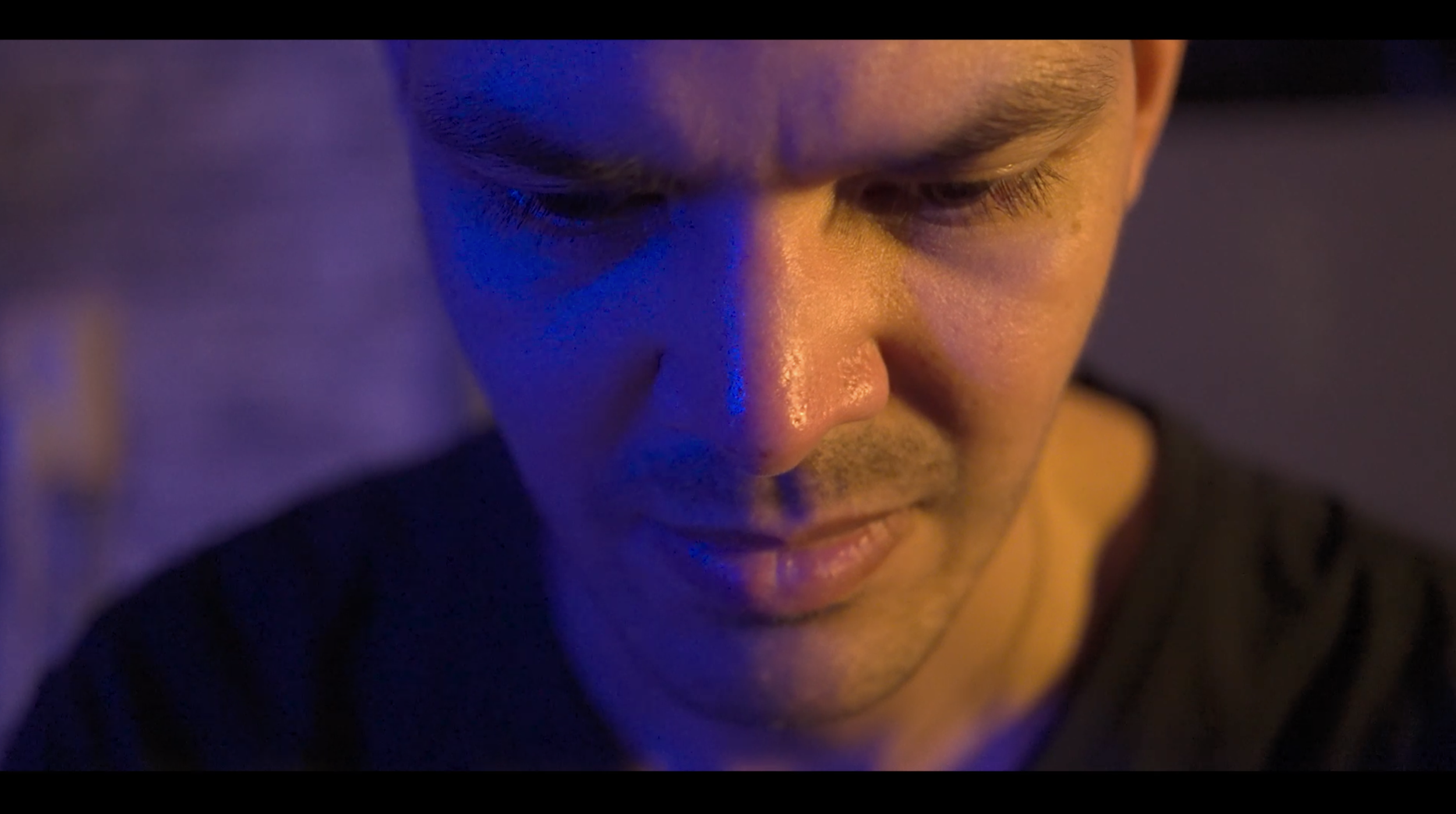 Close-up van een man met een rustige blik, licht op zijn gezicht en een blauwe en oranje gloed op de huid.